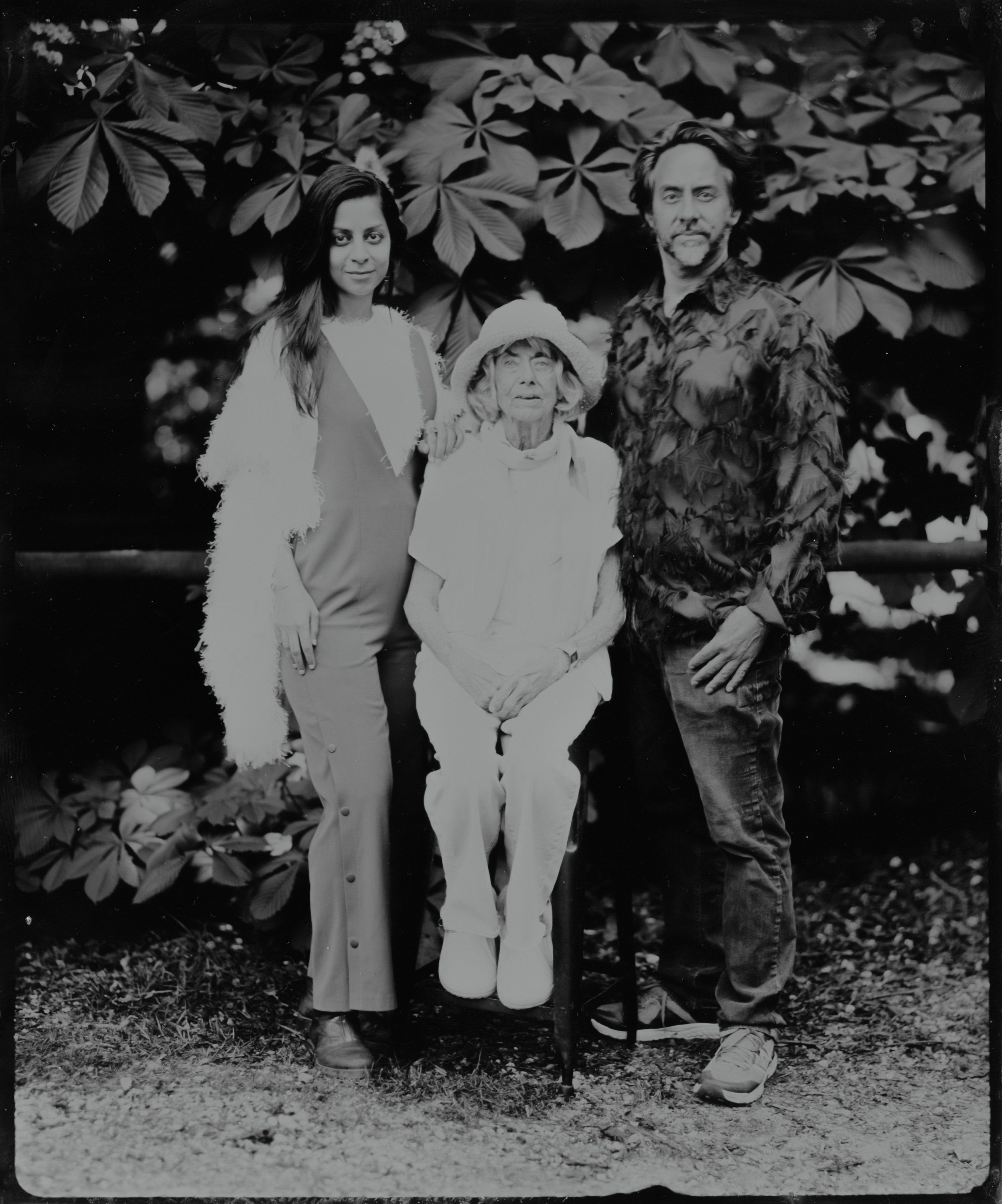 Black and white portrait of three people standing together among leafy foliage, soft natural light creating gentle shadows across their faces and clothing