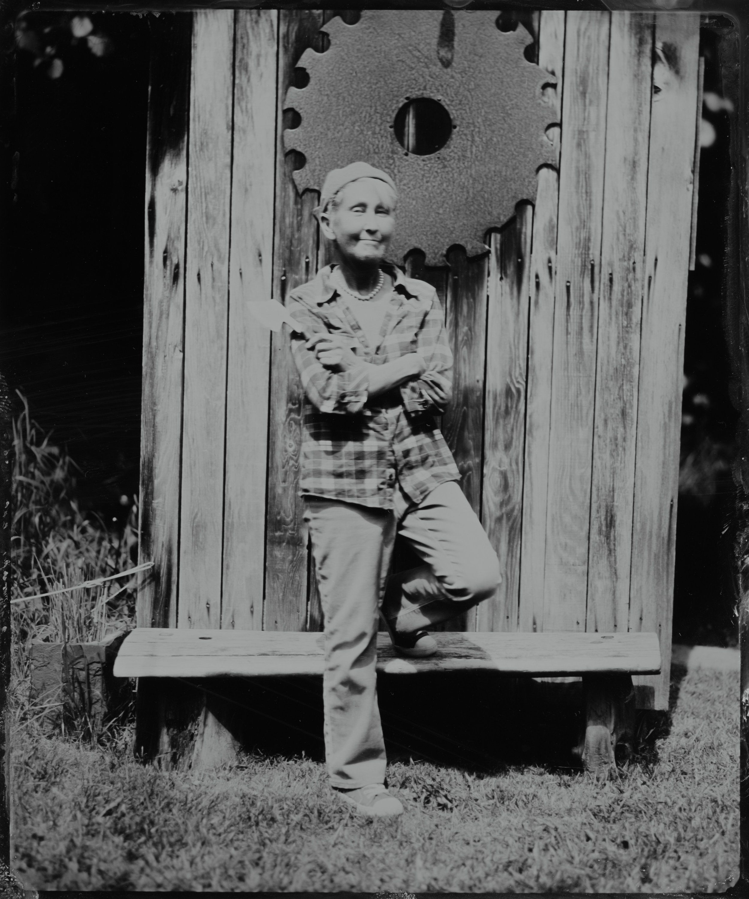 Man in plaid shirt leaning against rustic wooden outhouse door with circular cutout, black and white photograph
