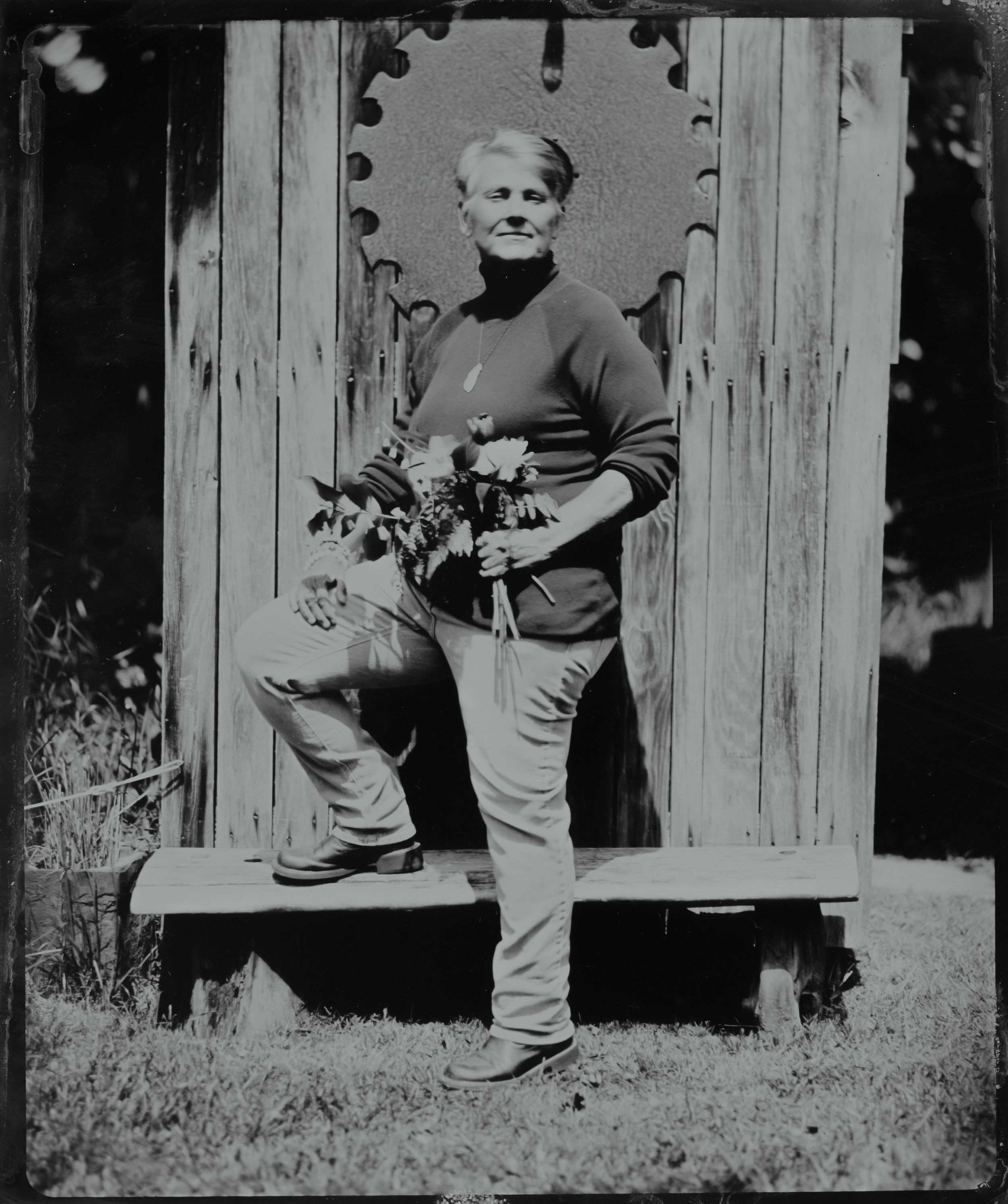 Elderly woman in vintage clothing holds wildflowers while standing on wooden steps, photographed in black and white with natural backlighting