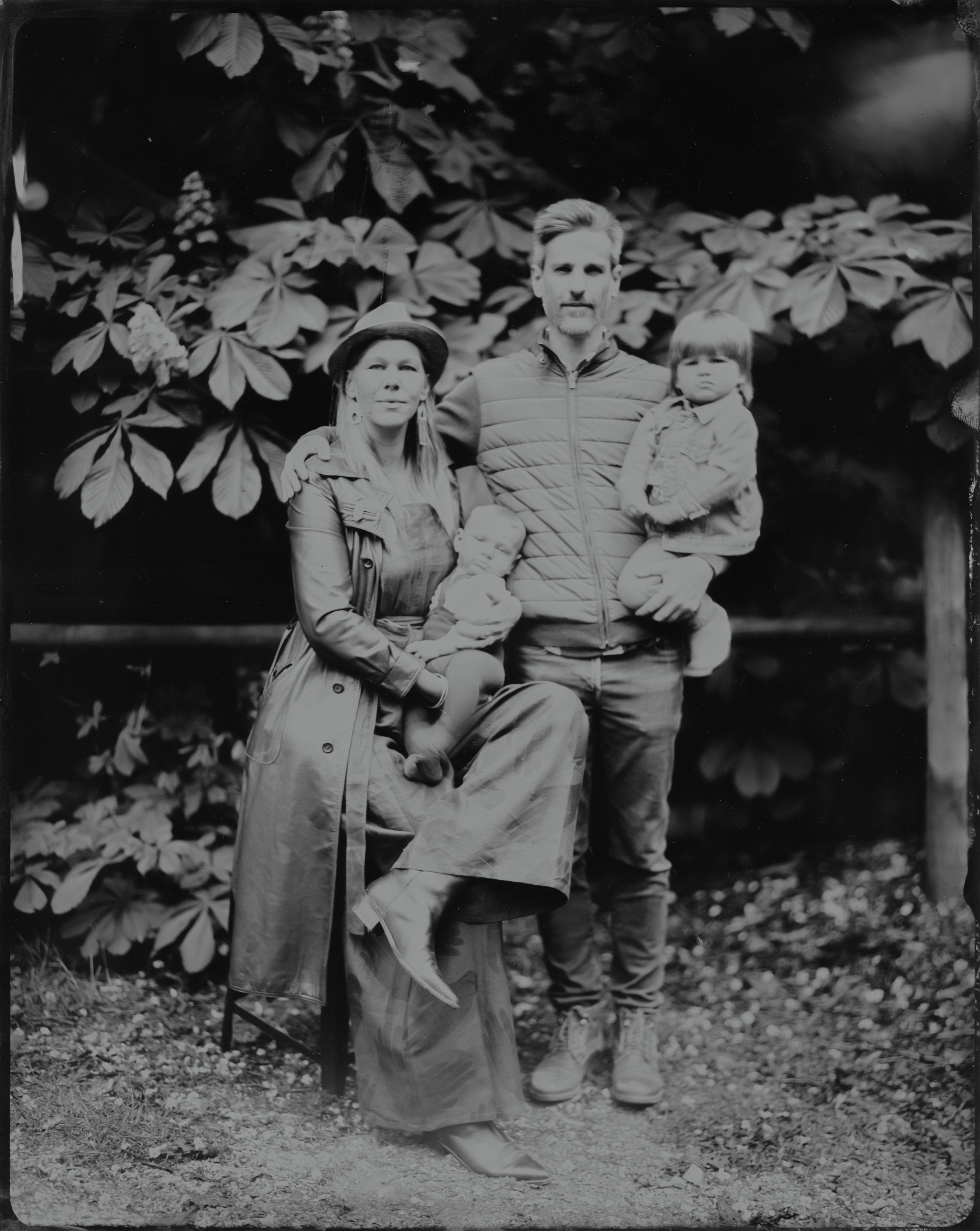 Family of three posed beneath leafy branches, soft natural light filtering through foliage in this intimate black and white portrait