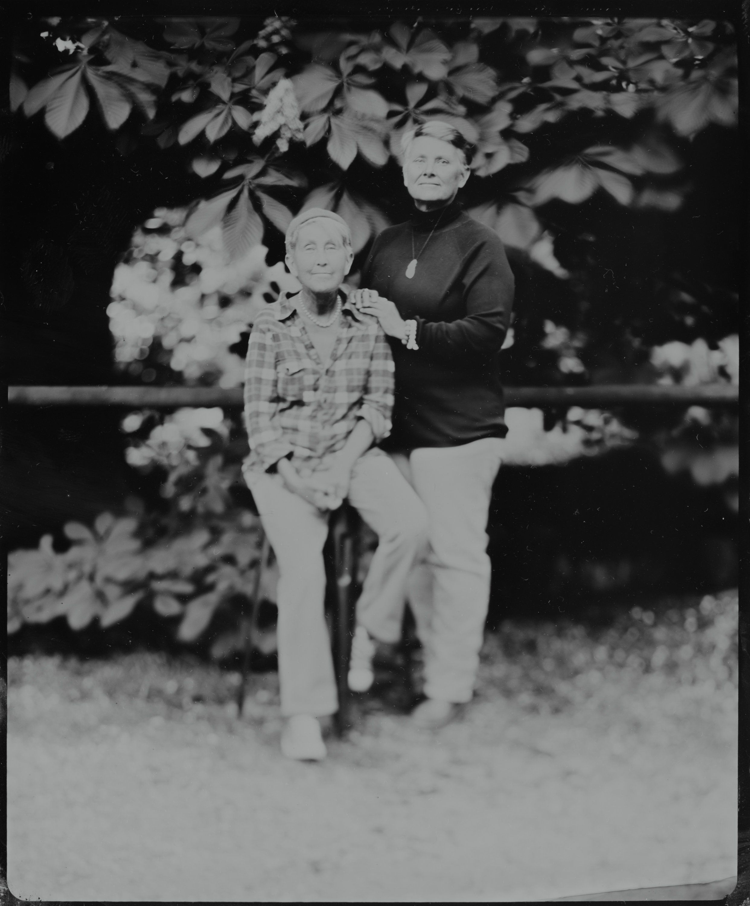 Two people stand together under flowering branches in dappled evening light, black and white photograph with soft, romantic mood