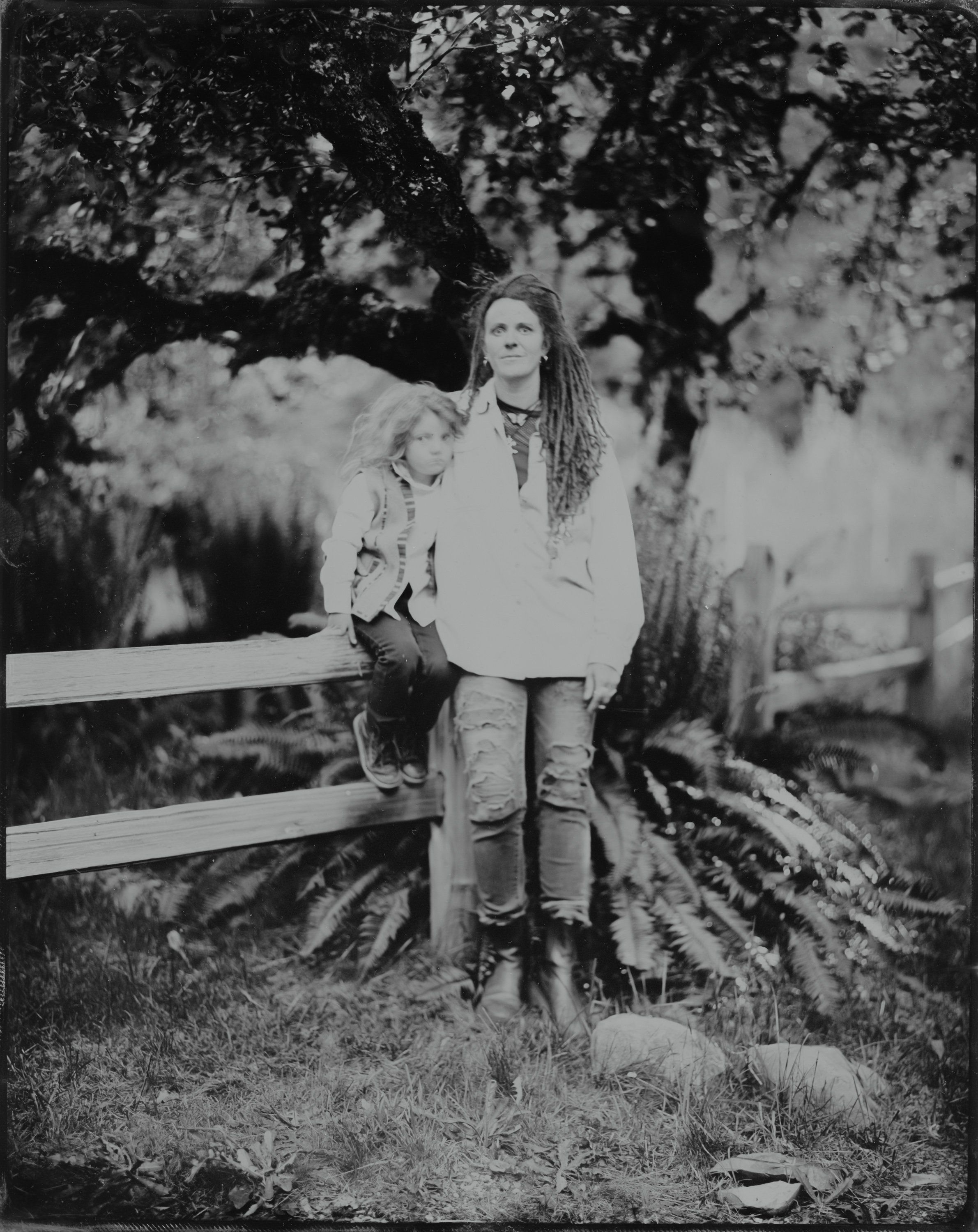 Child in white shirt leaning against wooden fence beneath leafy tree, black and white portrait with dappled shadow