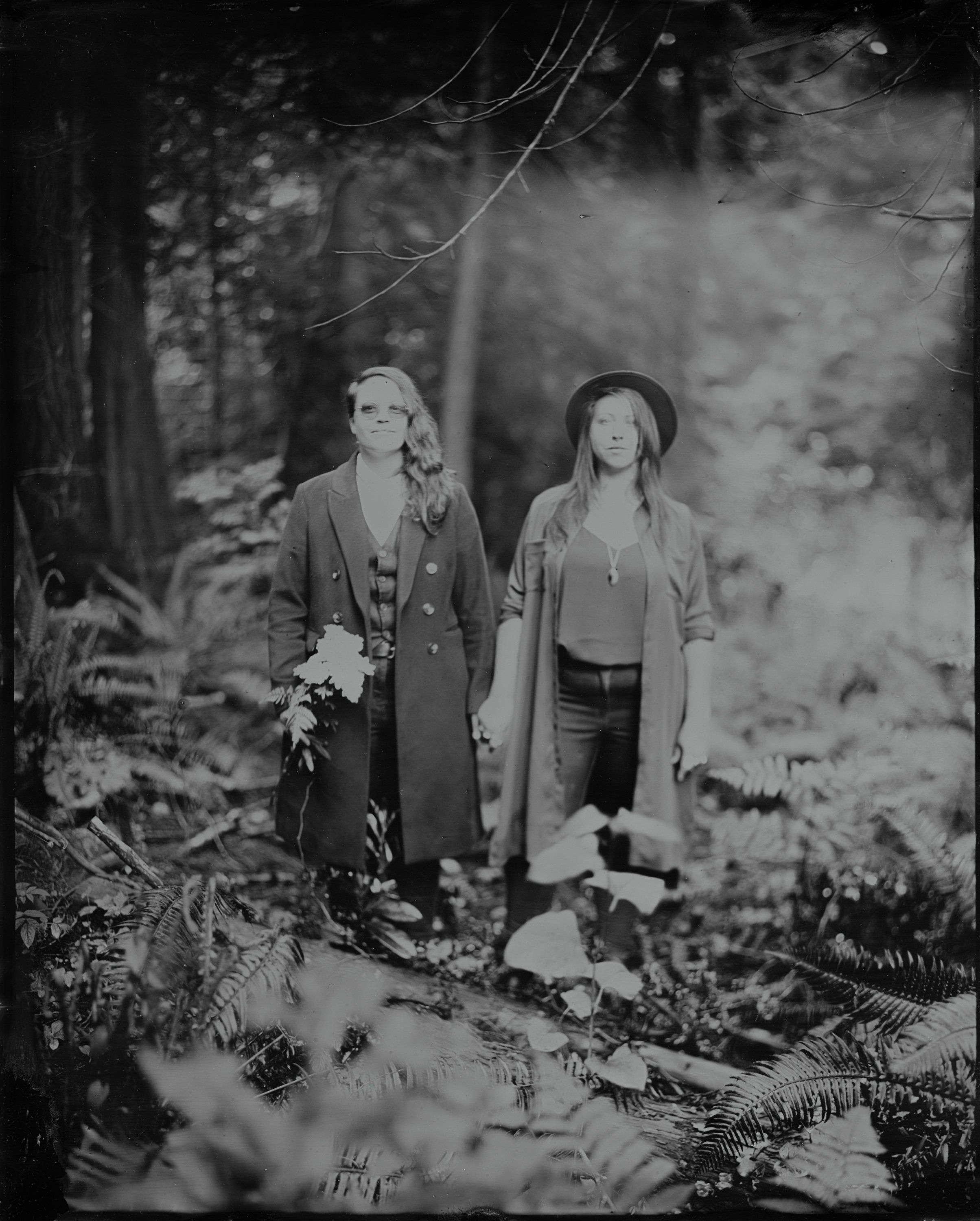 Two women in vintage coats stand among ferns in a misty forest, rendered in soft black and white
