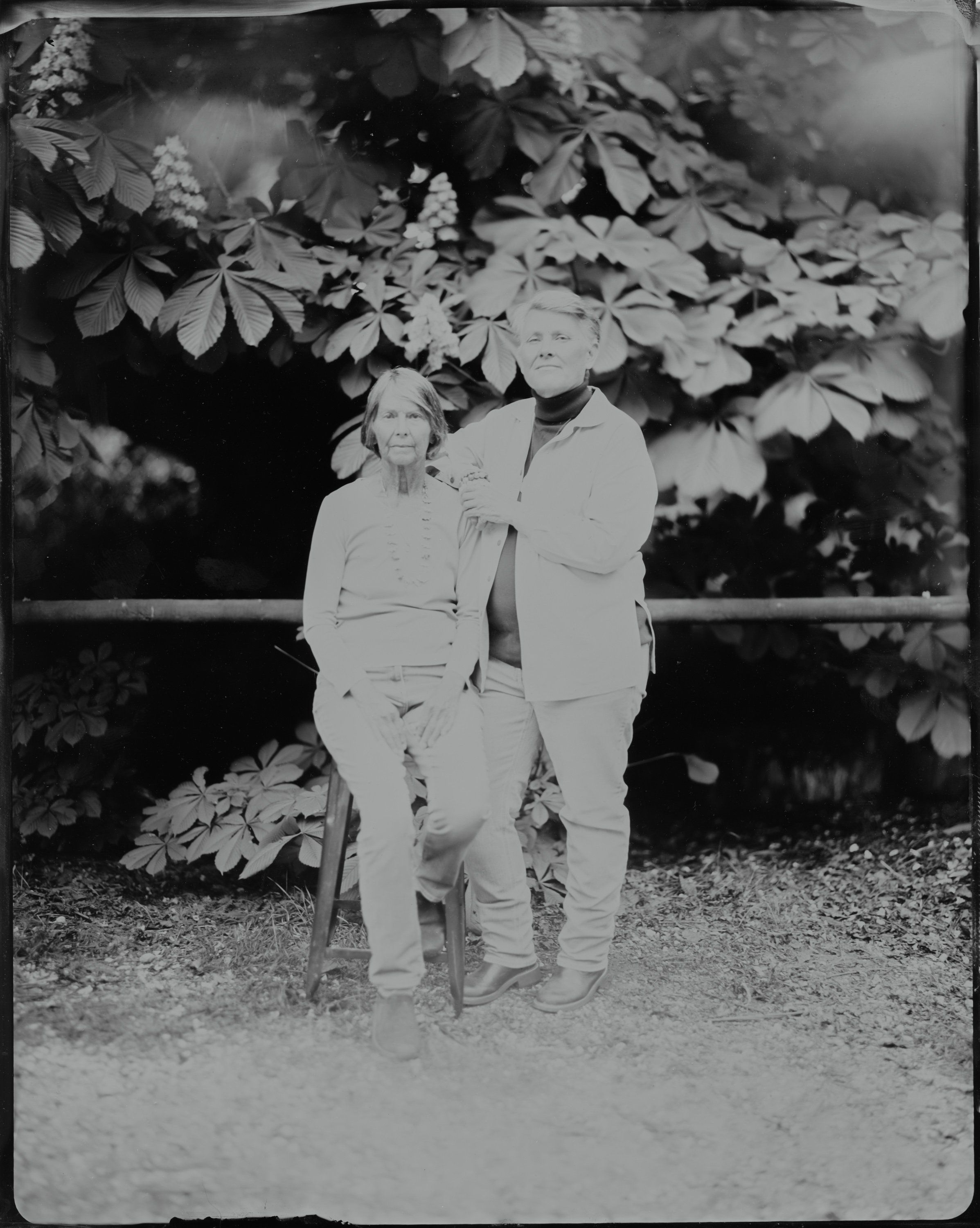 Couple in light suits standing together under leafy vines in a black and white portrait, soft garden setting