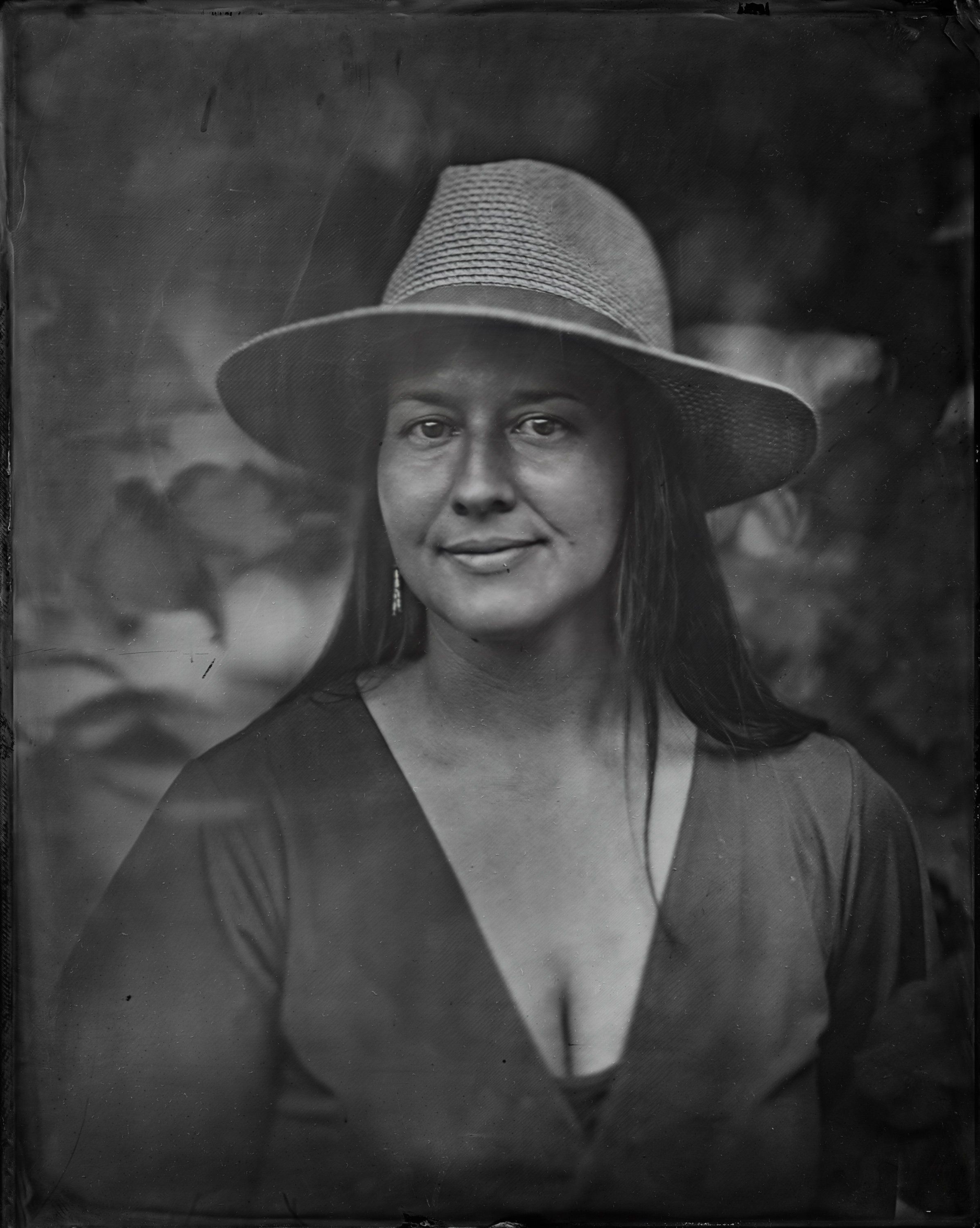 Woman in dark blouse and wide-brimmed hat gazes directly at camera with subtle smile in soft, diffused light