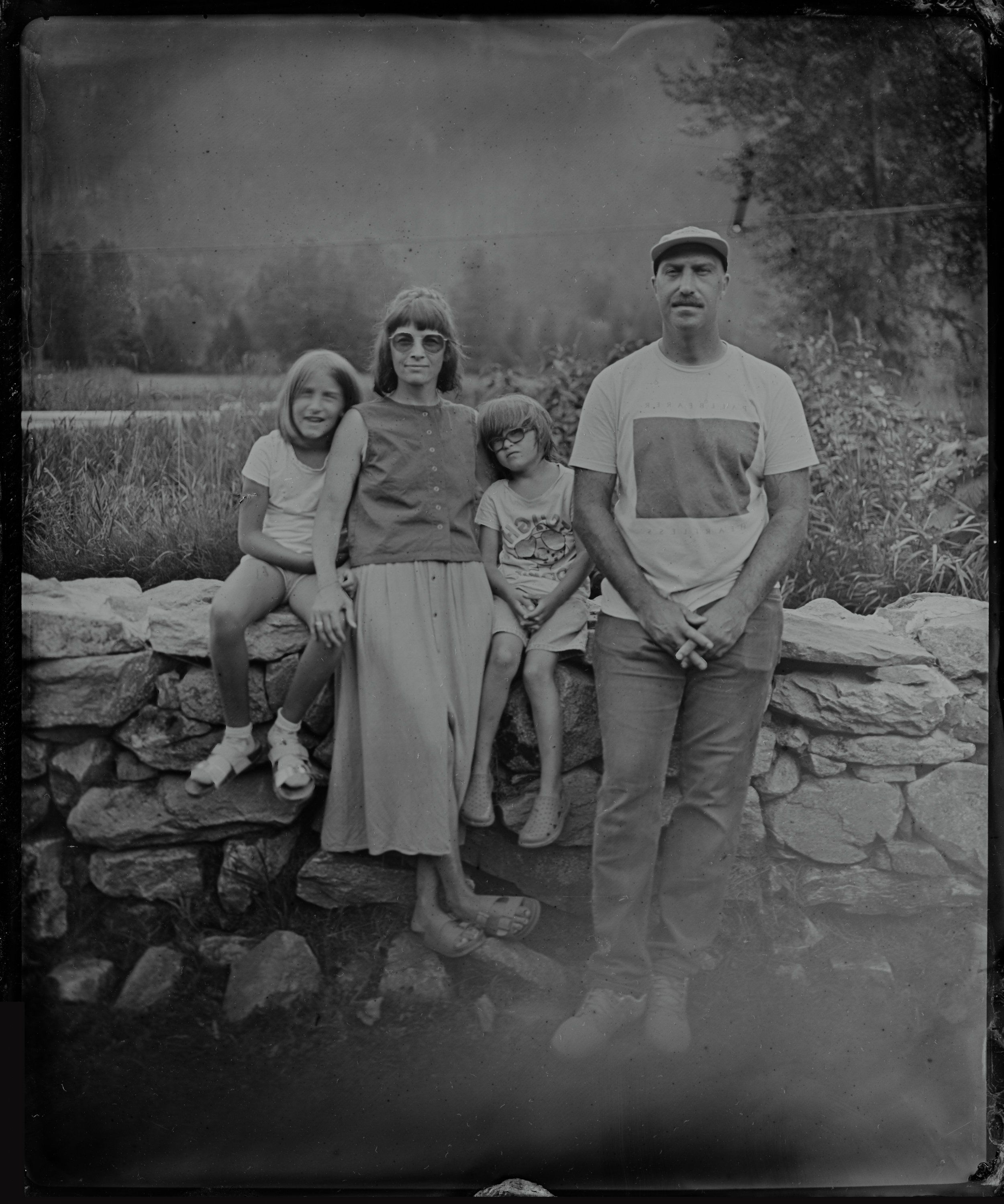Family of four standing together by a stone wall in a misty landscape, rendered in soft black and white tones