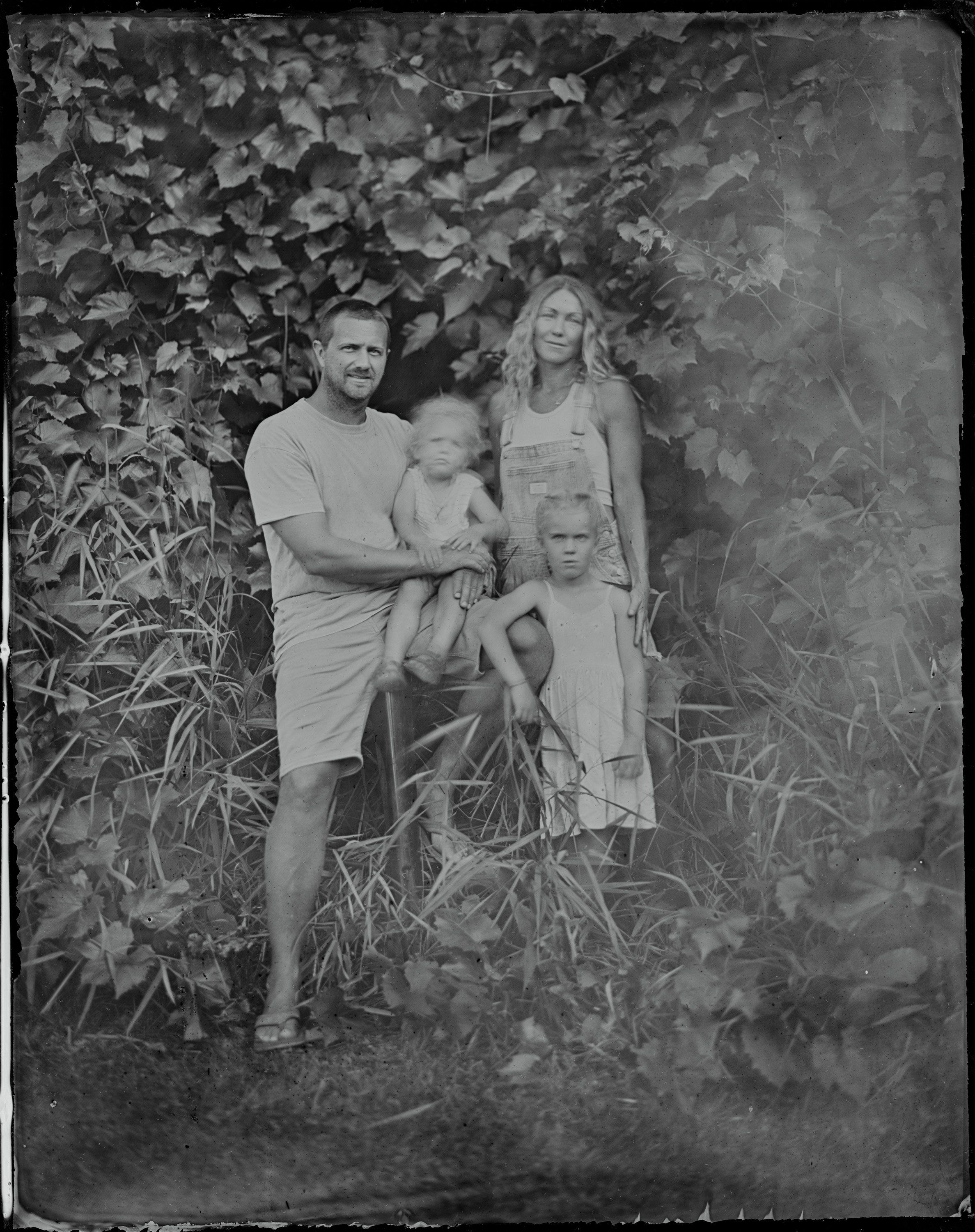 Family of three posed among overgrown vines against a weathered wall, soft natural light creating gentle shadows in this black and white portrait