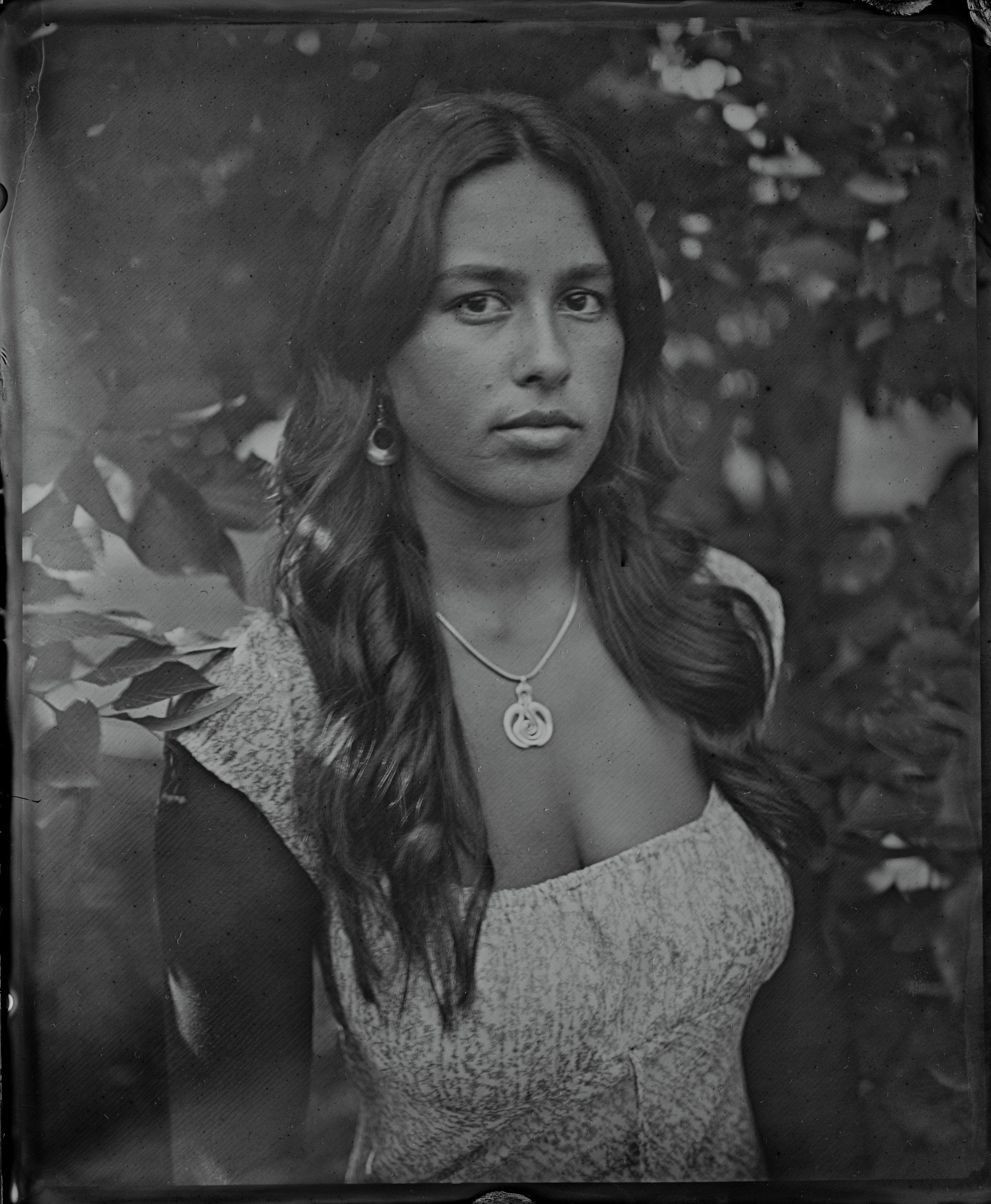 Woman with long dark hair and pendant necklace gazes directly at camera, surrounded by soft foliage in moody black and white tintype portrait