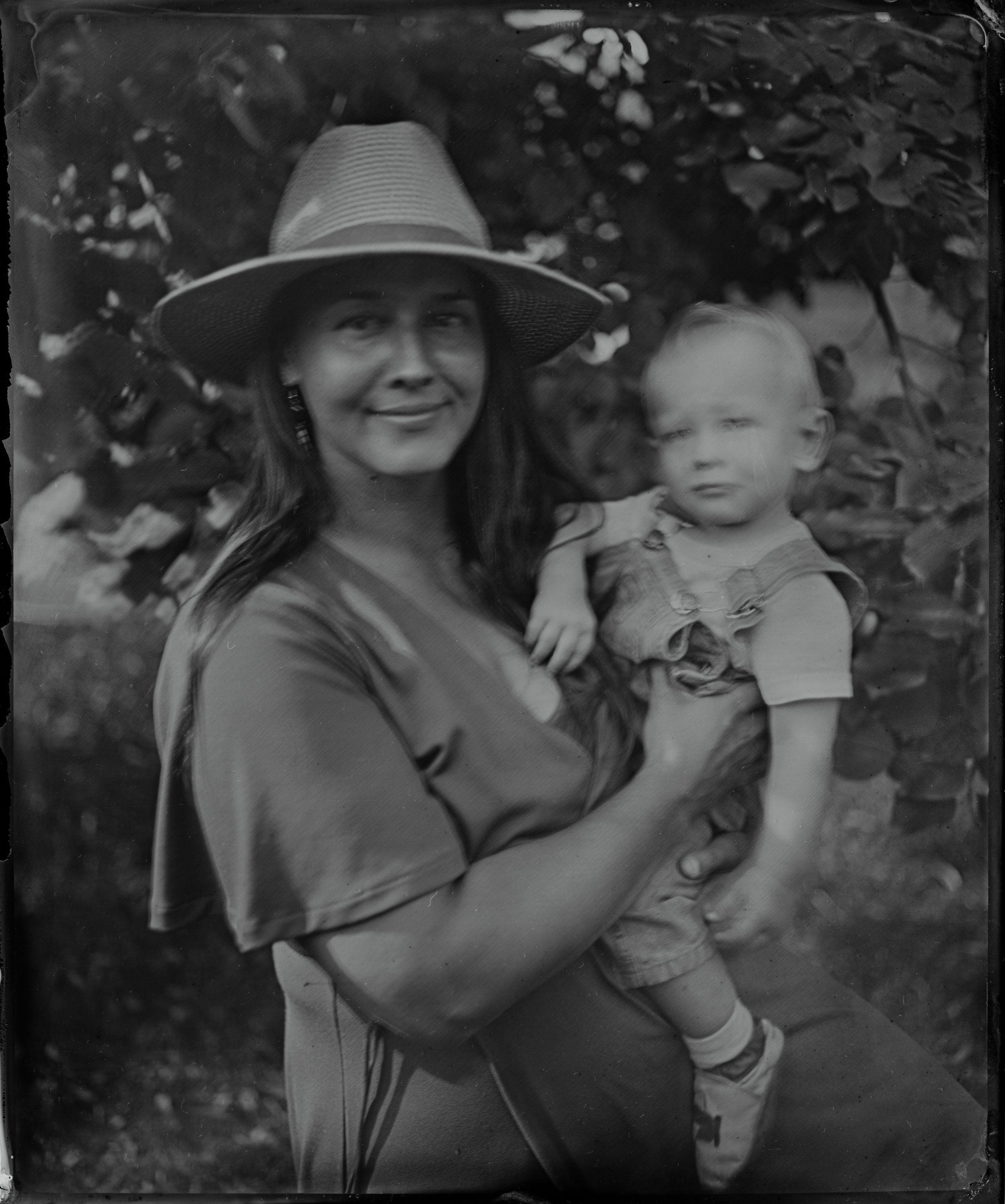 Woman in wide-brimmed hat holding young child outdoors, dappled light through foliage, warm intimate portrait