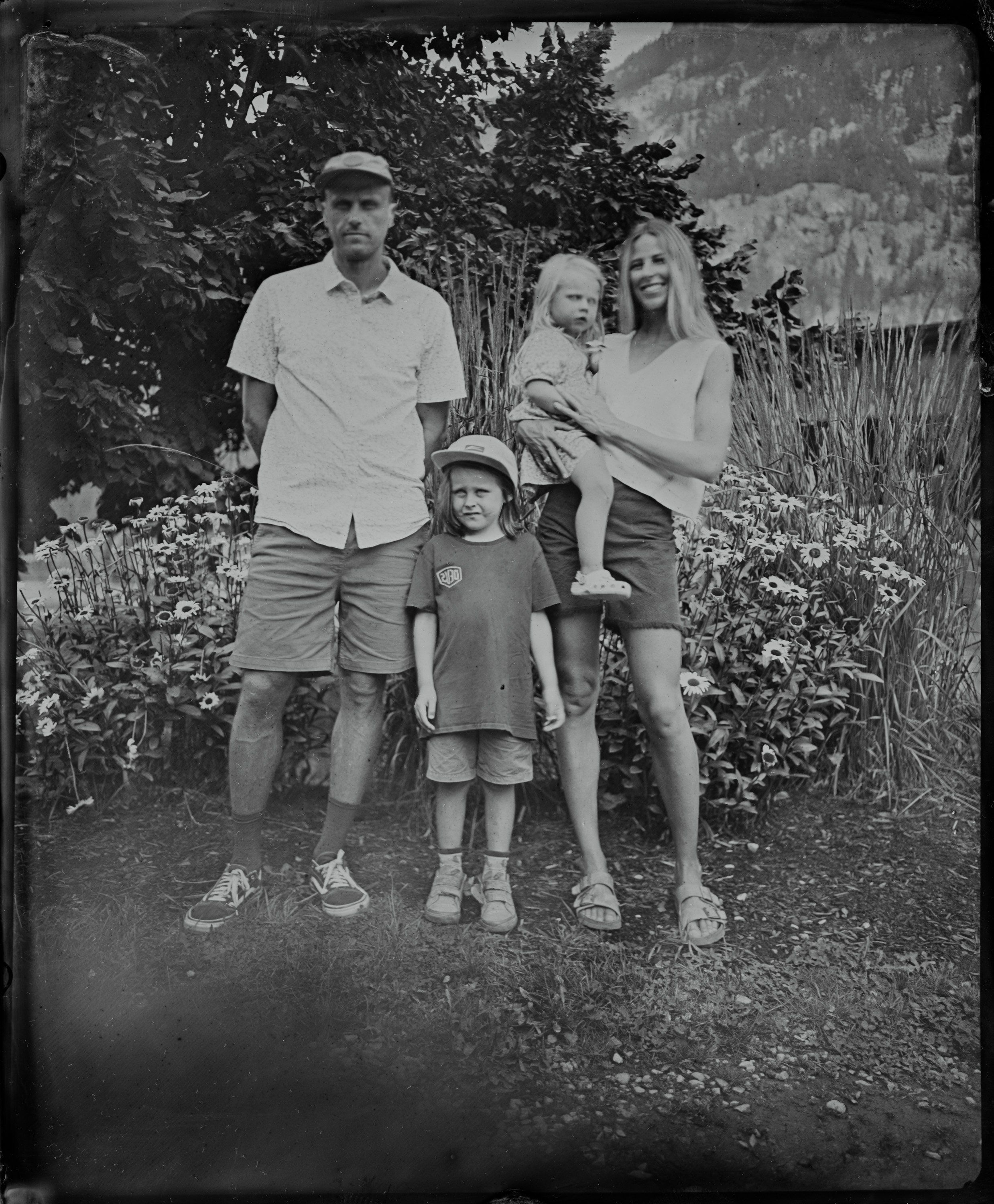 Family of four stands in a garden with mountains behind, dressed in light summer clothing, rendered in soft black and white tones