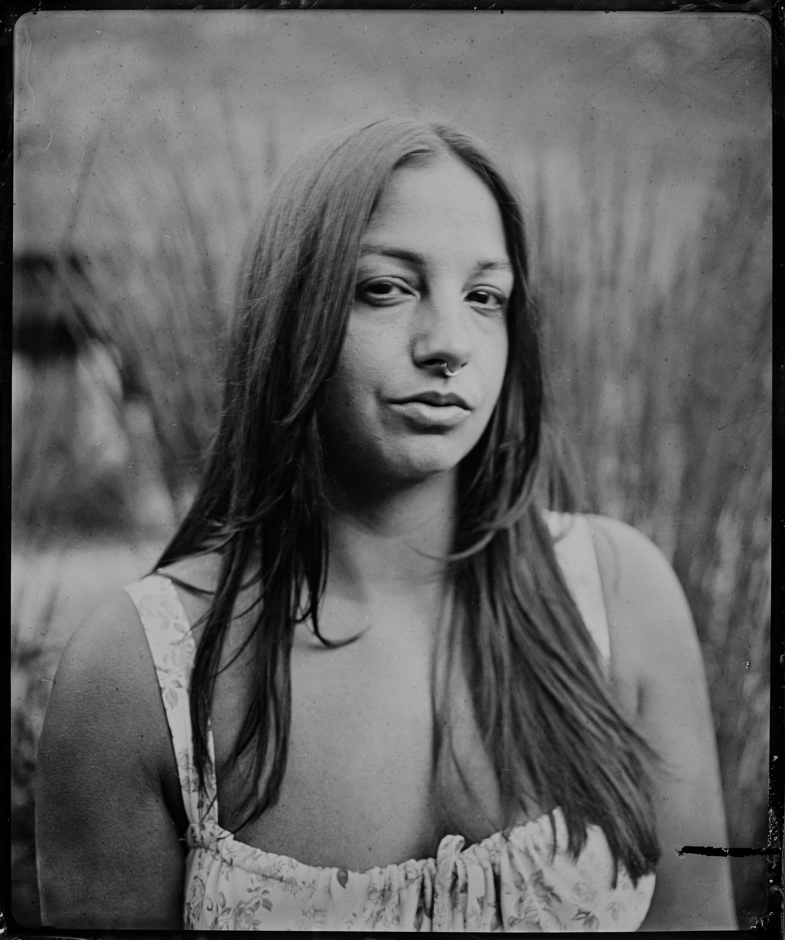 Woman with long dark hair gazes pensively aside in soft-focus portrait, rendered in classic tintype aesthetic against blurred natural background