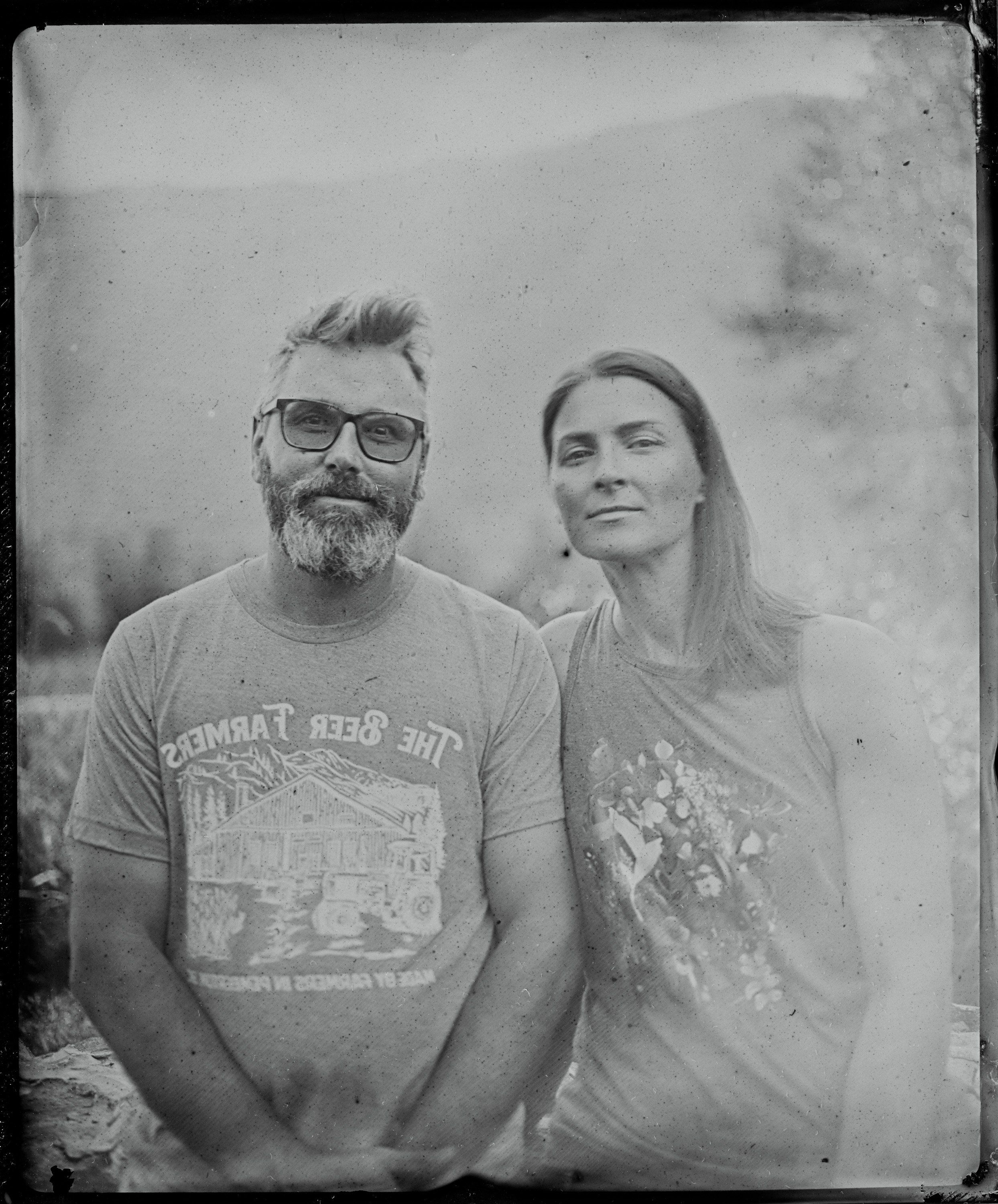 Two people stand together against a softly blurred background in this black and white tintype portrait, their expressions calm and direct