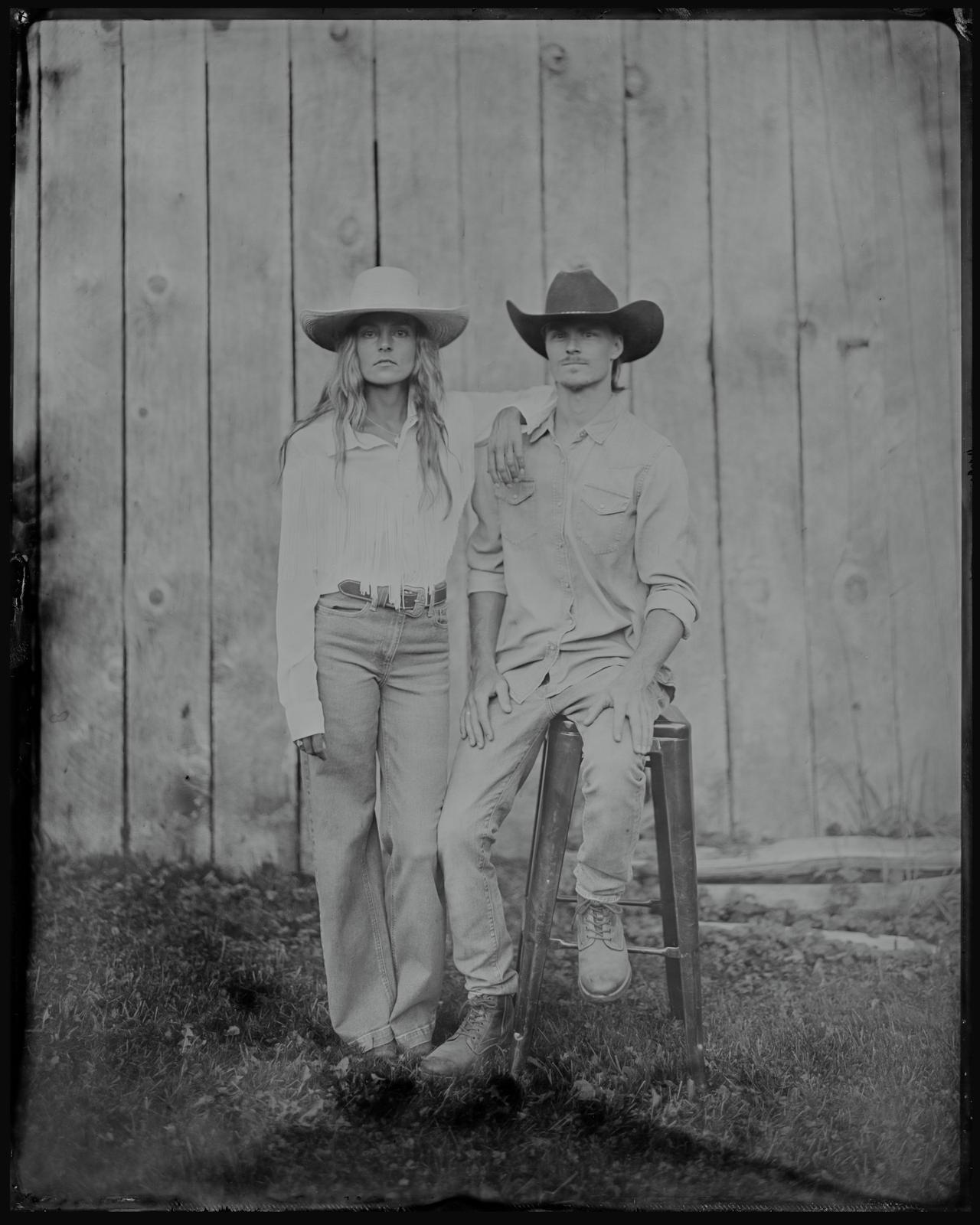 Two people in cowboy hats stand shoulder to shoulder against a plain wall, gazing upward with contemplative expressions