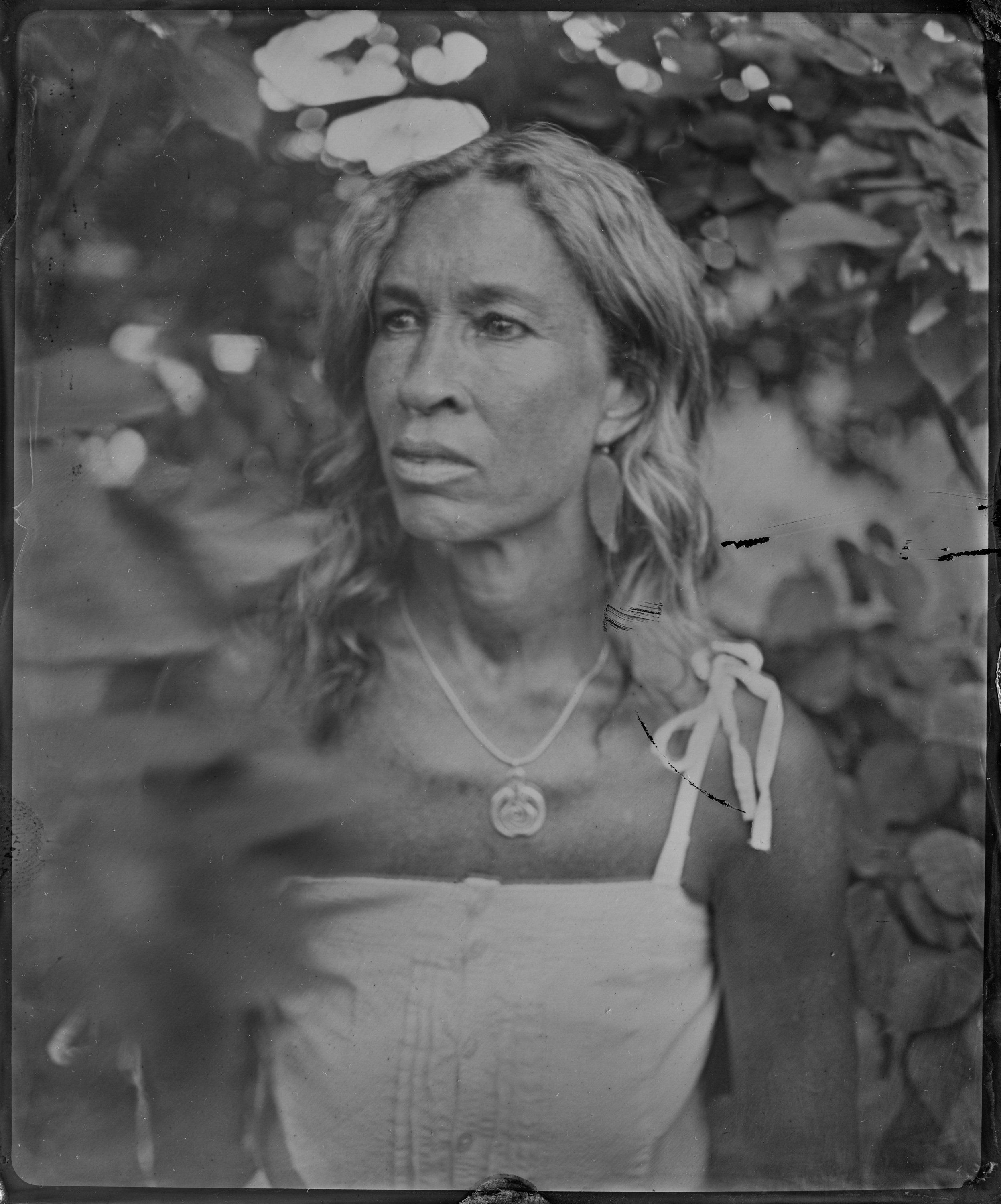 Woman with long hair and pendant necklace gazes thoughtfully amid soft, dappled foliage in monochrome light
