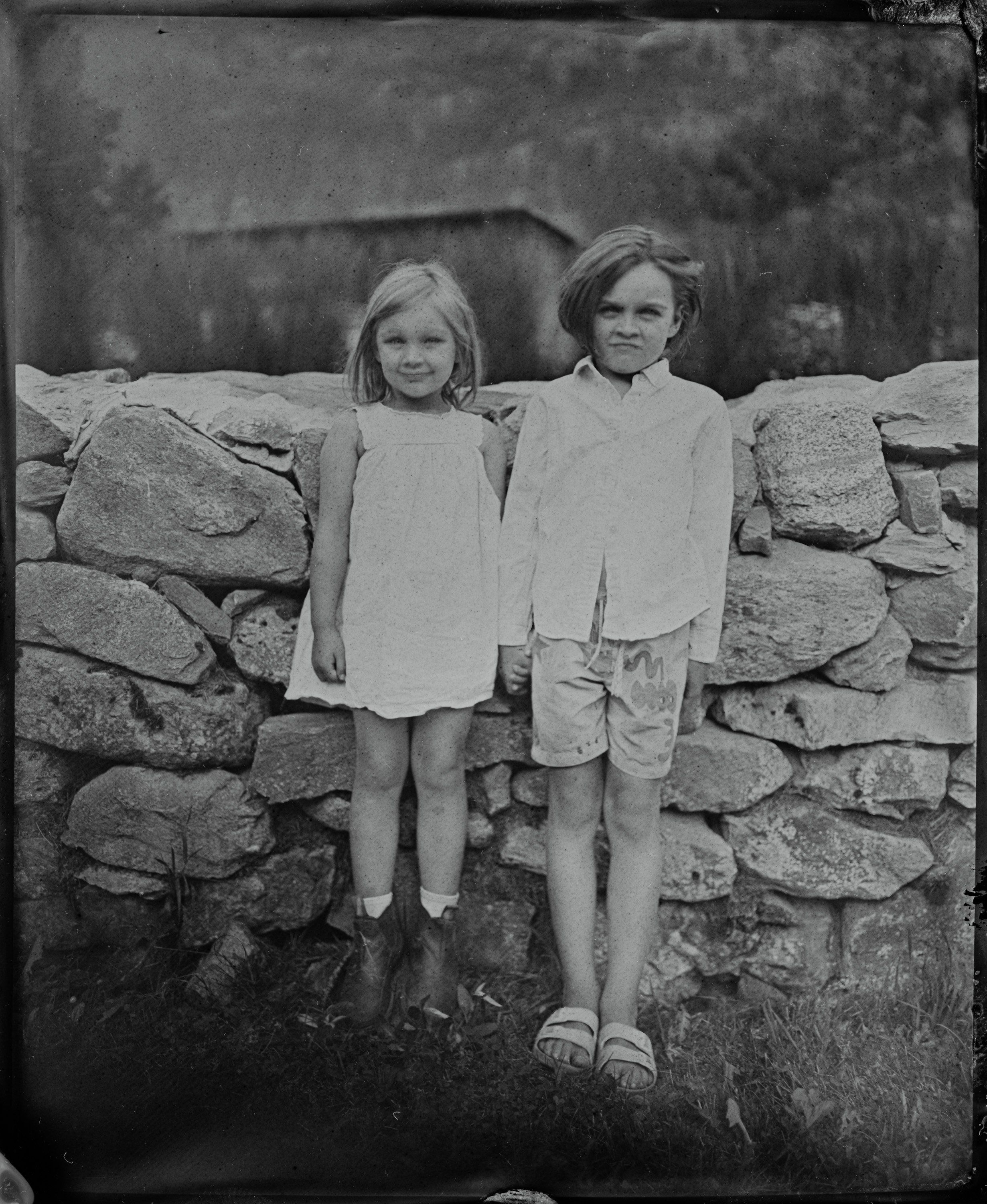 Two young children stand barefoot against a weathered stone wall, wearing simple light-colored clothing in this soft-focus black and white portrait