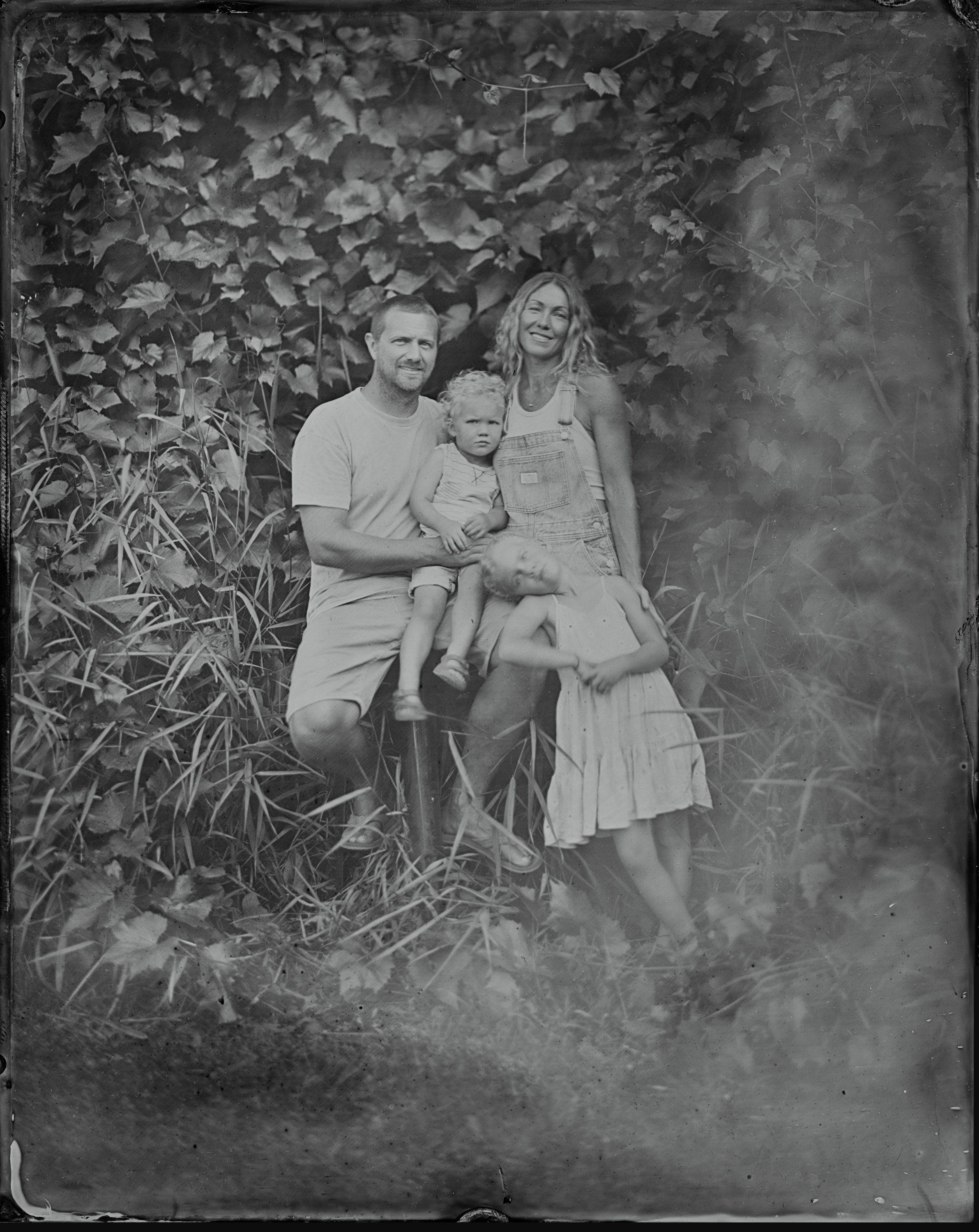 Man, woman, and young child posed together among ivy-covered vines in soft natural light, black and white tintype portrait