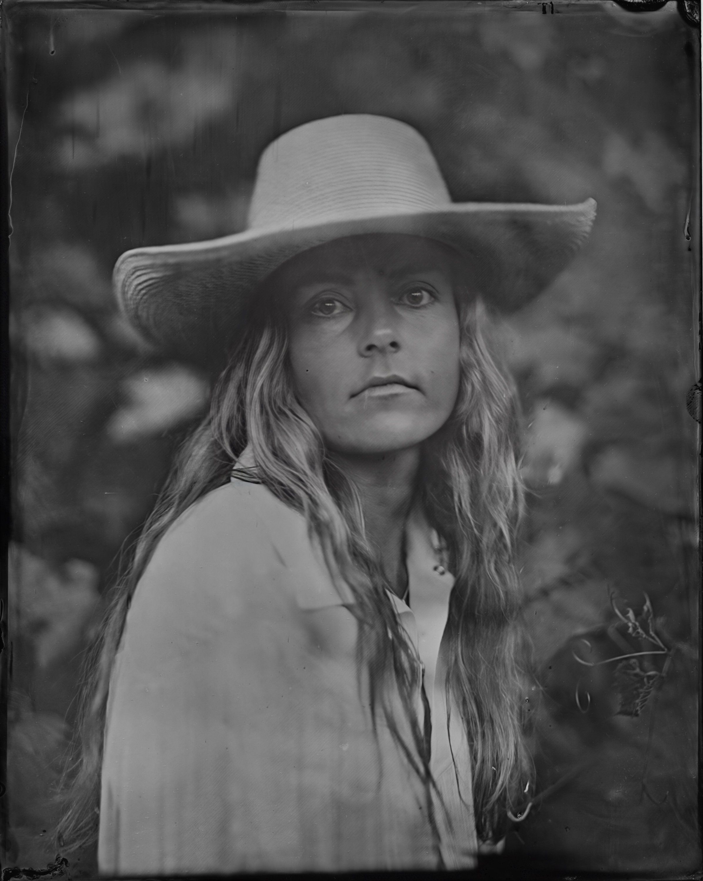 Portrait of a woman with long wavy hair wearing a wide-brimmed cowboy hat and light-colored shirt, gazing over her shoulder against a soft, blurred natural b…