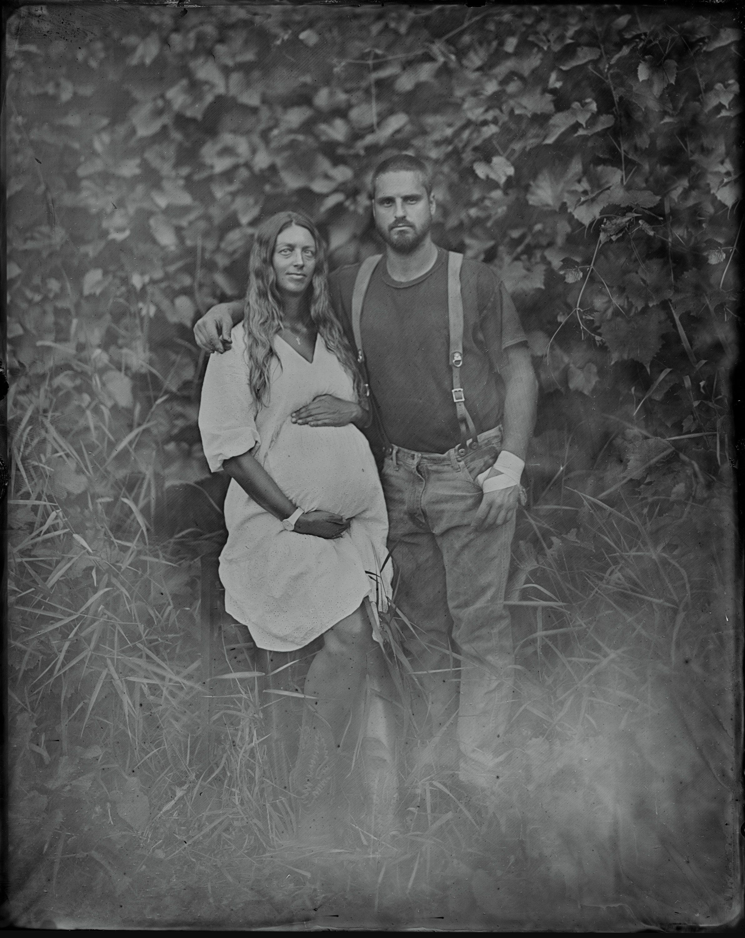 Pregnant woman in white dress stands between two men against overgrown foliage backdrop in moody tintype style portrait