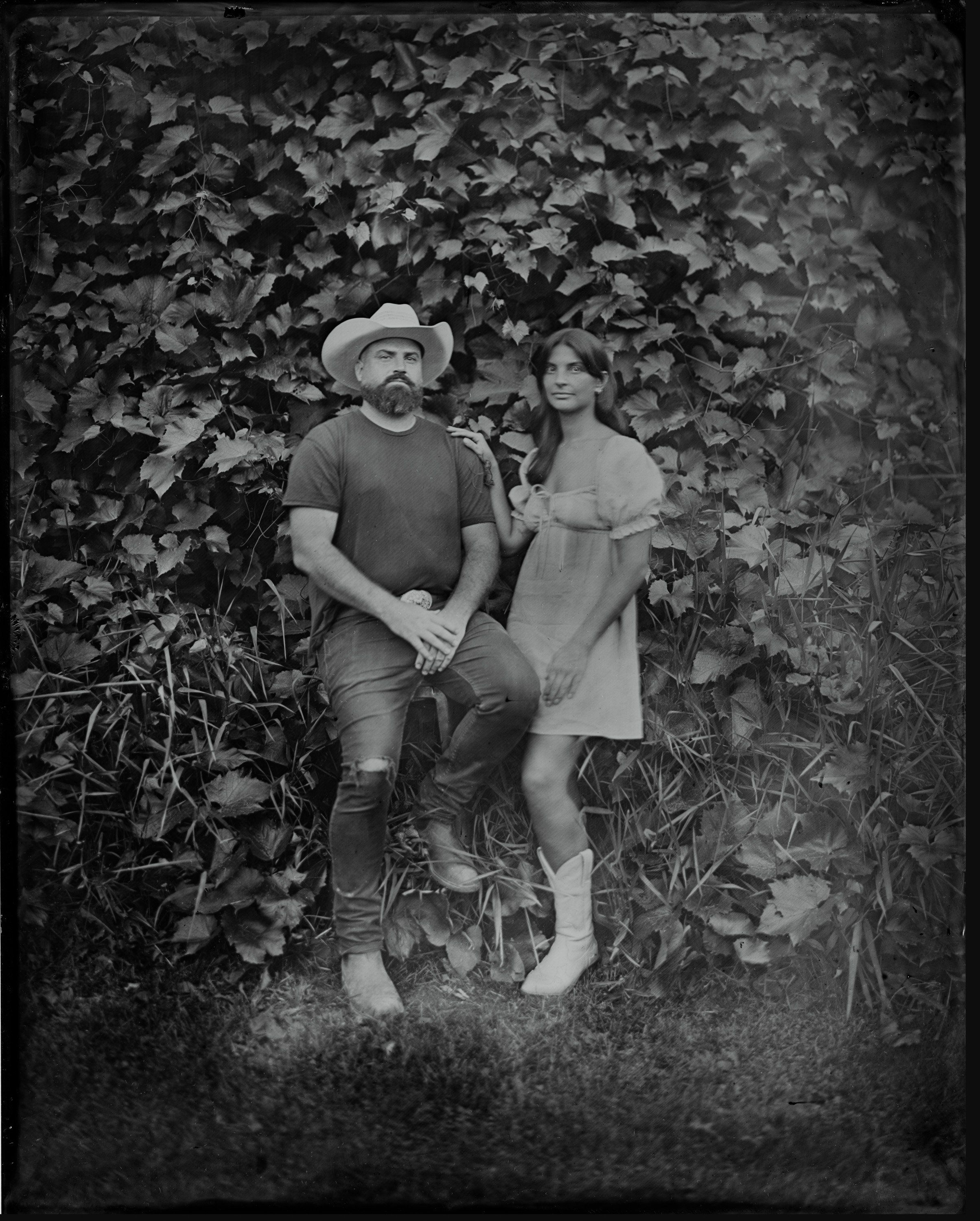Man in cowboy hat and woman in light dress recline against ivy-covered wall in black and white tintype