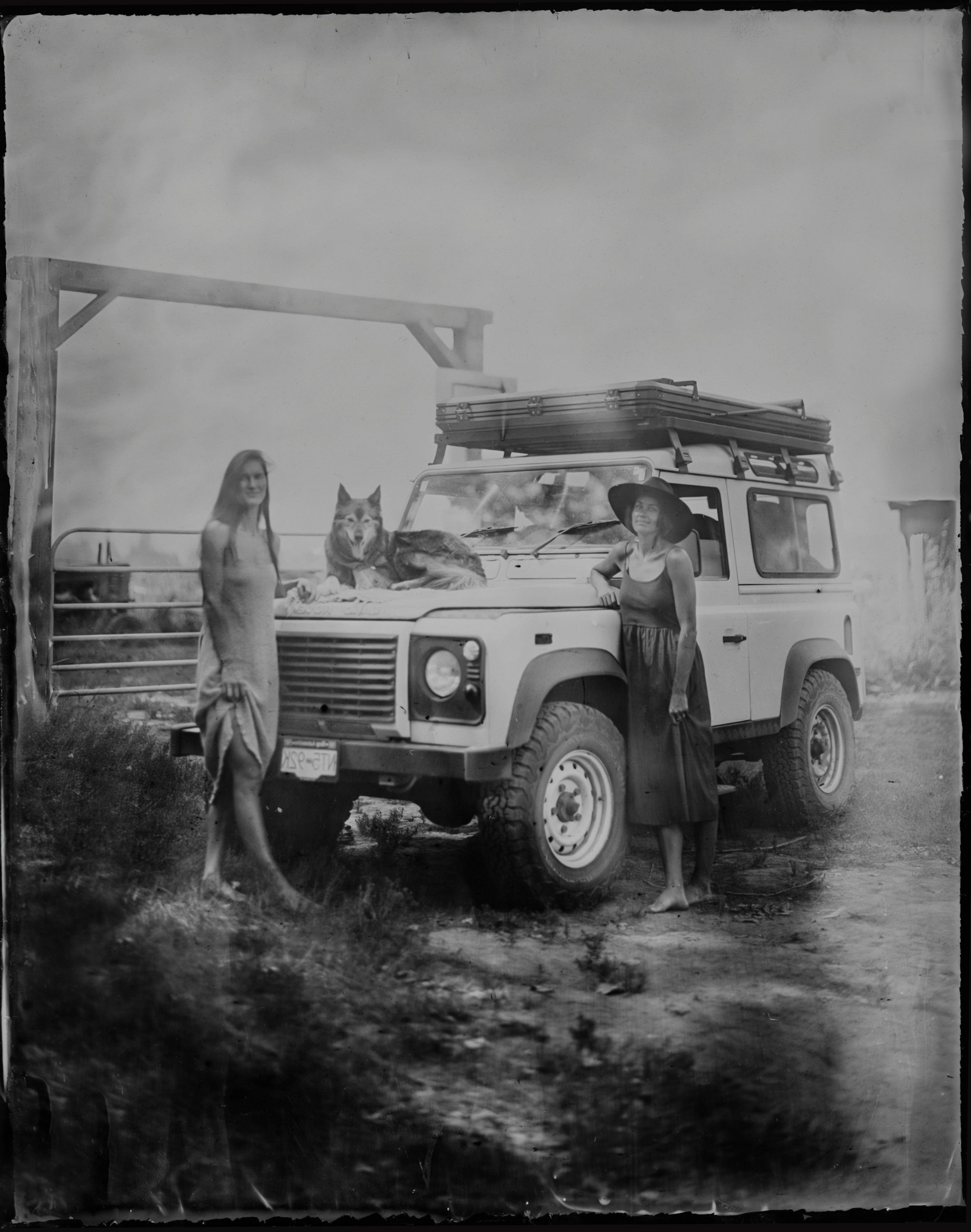 Two women stand beside a vintage Land Rover laden with gear, rain-soaked ground and overcast sky creating moody atmosphere