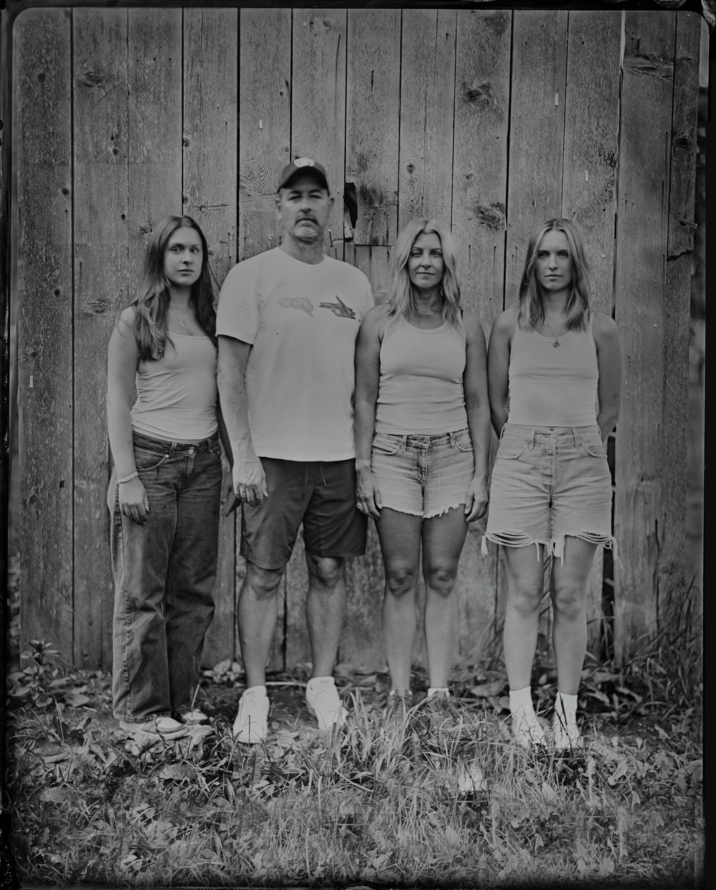 Four people stand against a weathered wooden wall in black and white, their casual poses and worn clothing creating an intimate, documentary-like portrait