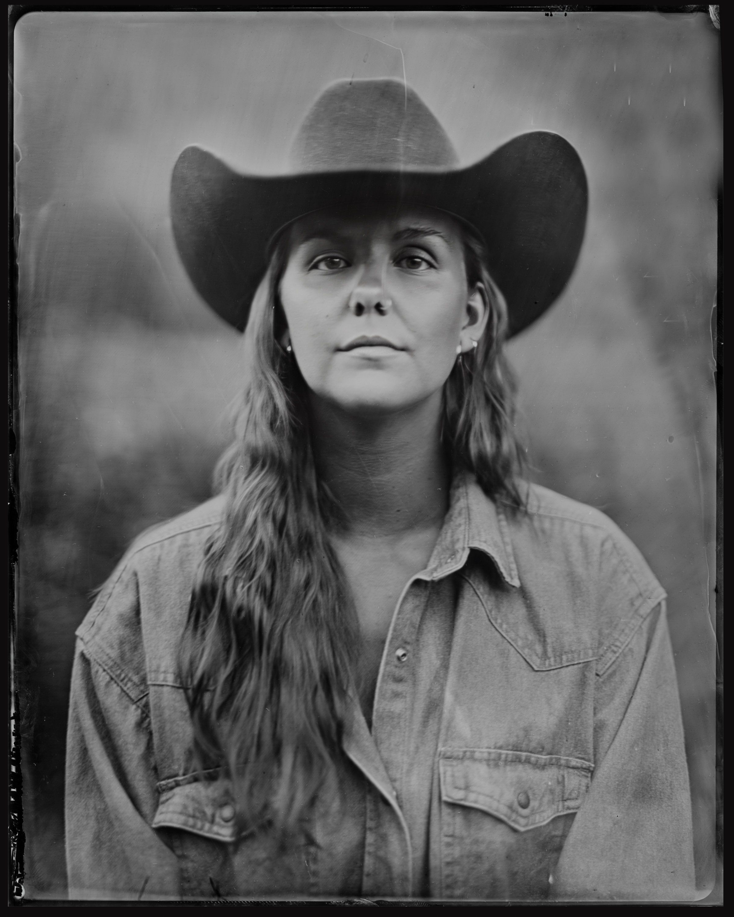 Portrait of a person with long hair wearing a cowboy hat and denim shirt, gazing upward with a contemplative expression in soft, diffused light