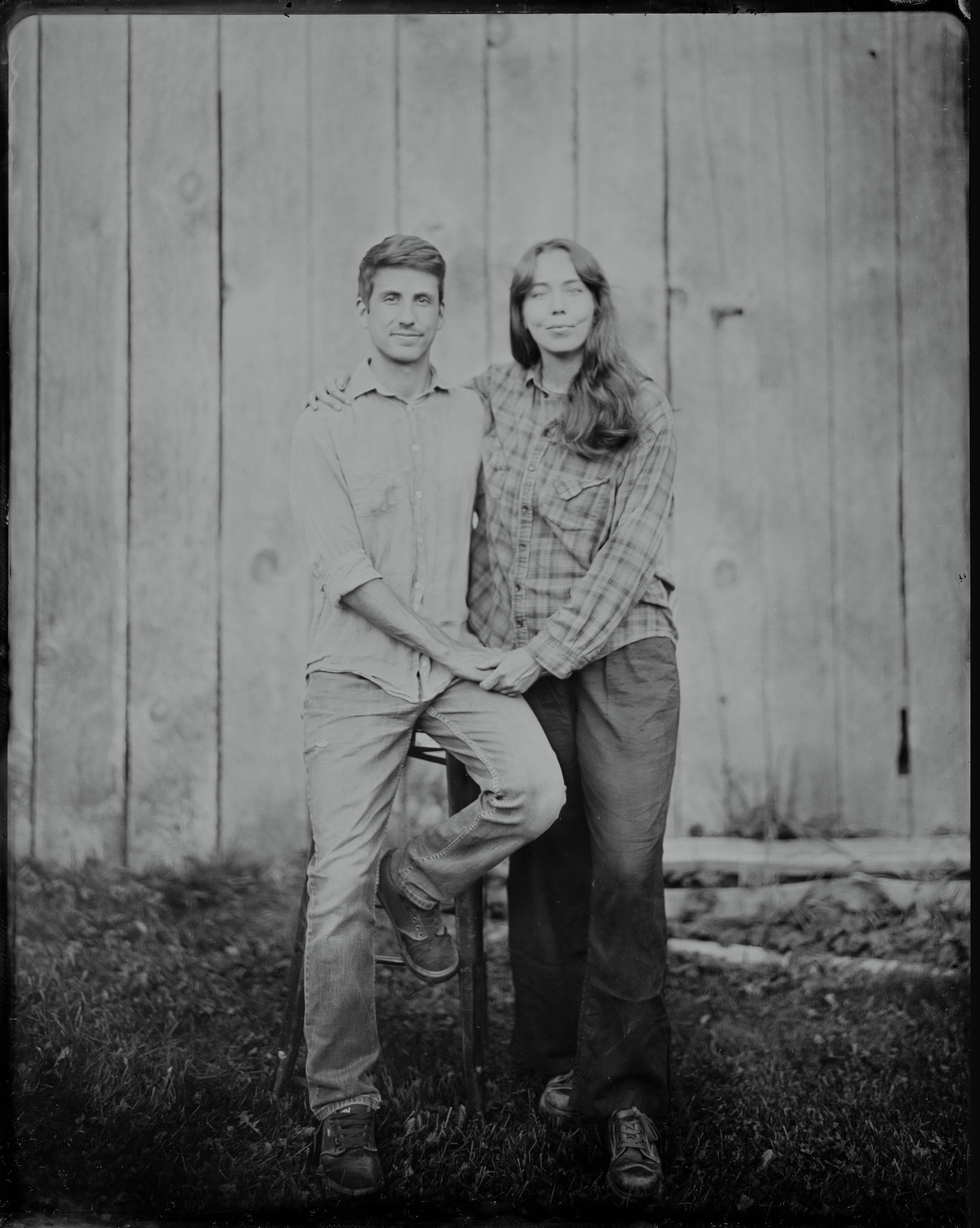 Two people in casual flannel stand against weathered wooden boards, holding a baseball bat between them in this black and white portrait