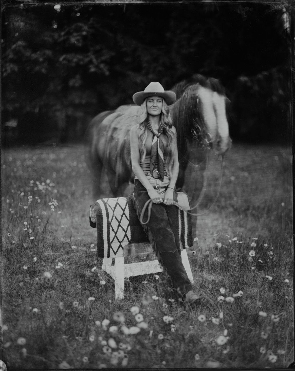 Woman in cowboy hat and fringe jacket sits with saddled horse in wildflower meadow, rendered in moody black and white tintype style