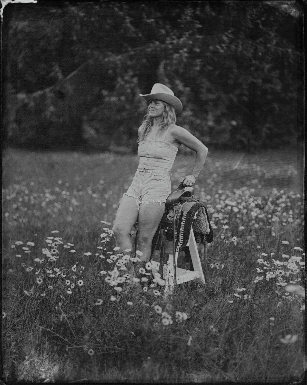 Cowgirl in white hat and denim shorts holds saddle in wildflower meadow, tintype portrait