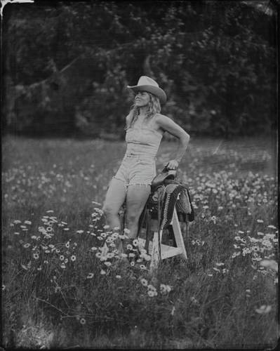 Cowgirl in white hat and denim shorts holds saddle in wildflower meadow, tintype portrait