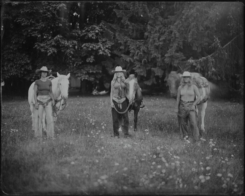 Cowgirl in a wide-brimmed hat holds a coiled rope while standing with her horse in a wildflower meadow, rendered in moody black-and-white tintype