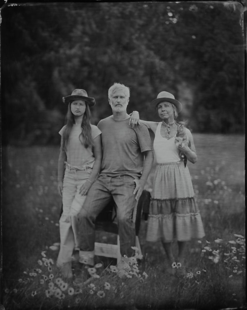 Three people in casual attire with hats stand shoulder-to-shoulder outdoors, framed by soft foliage and wildflowers in a tintype portrait