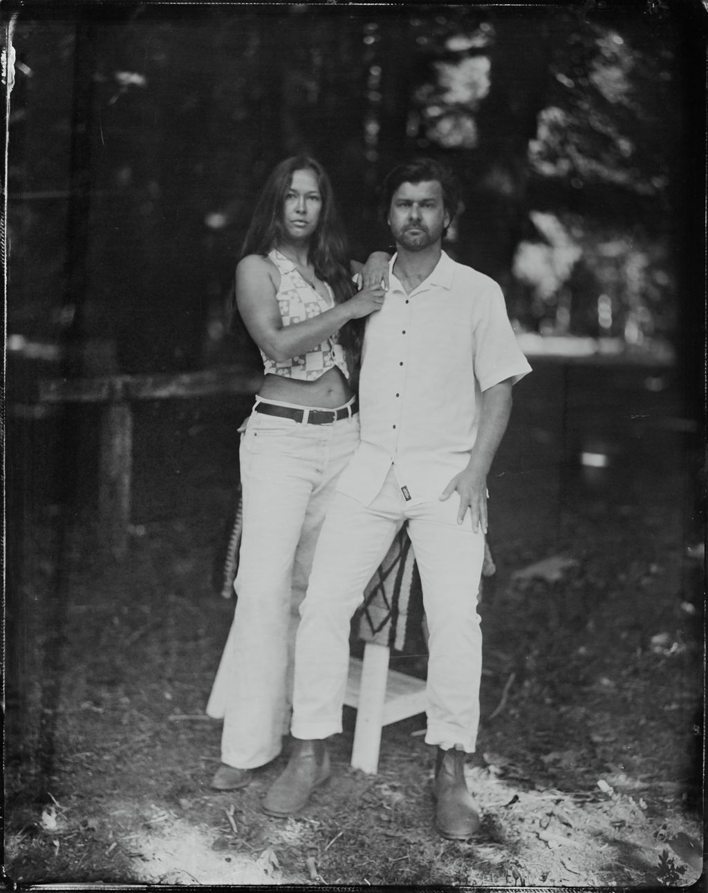 Couple in white clothing pose together at dusk near forest in wet-plate tintype style portrait, her hand on his shoulder with soft, dreamy bokeh lighting
