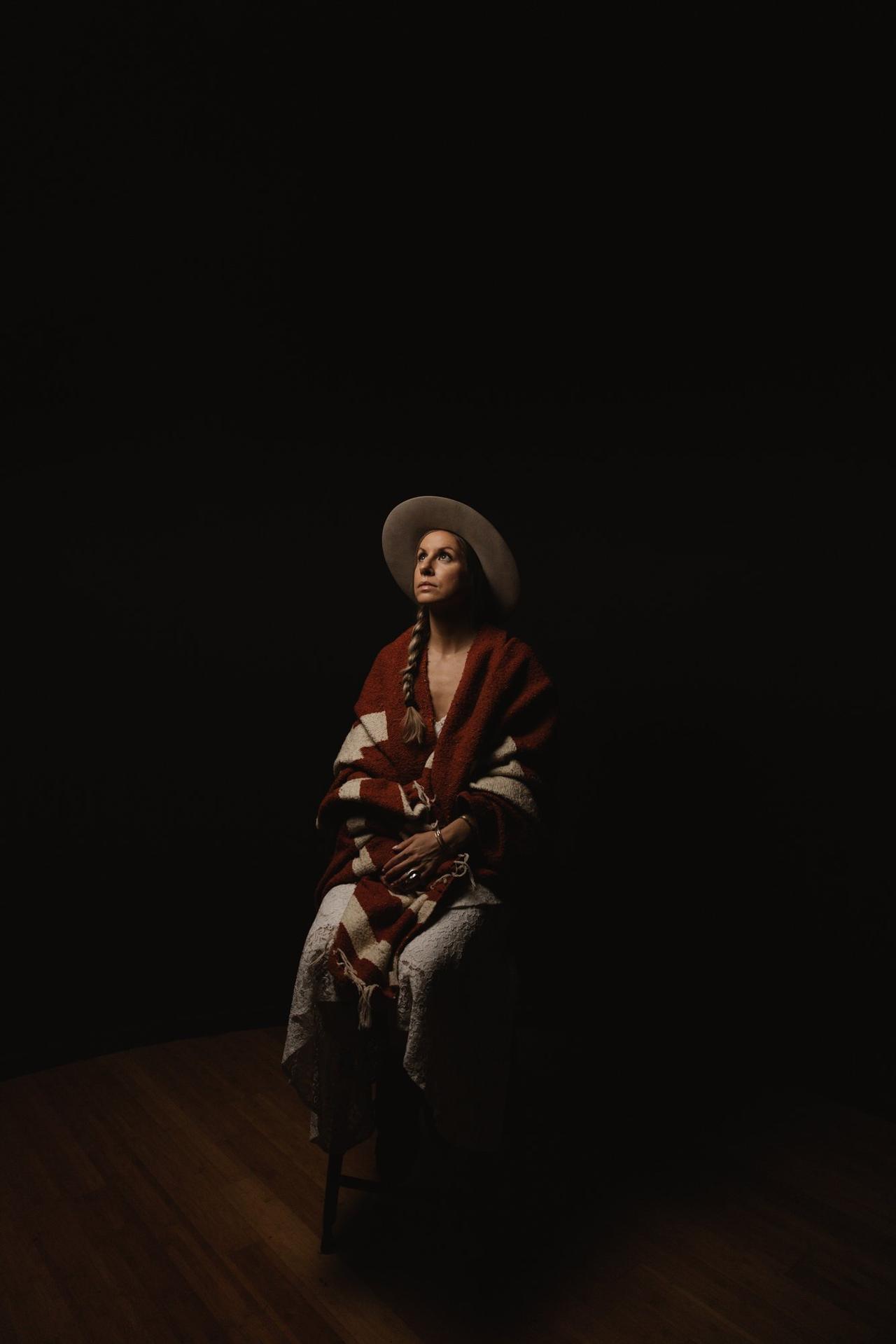 Woman in red and cream striped blanket and wide-brimmed hat gazes upward against black background, hands folded in lap