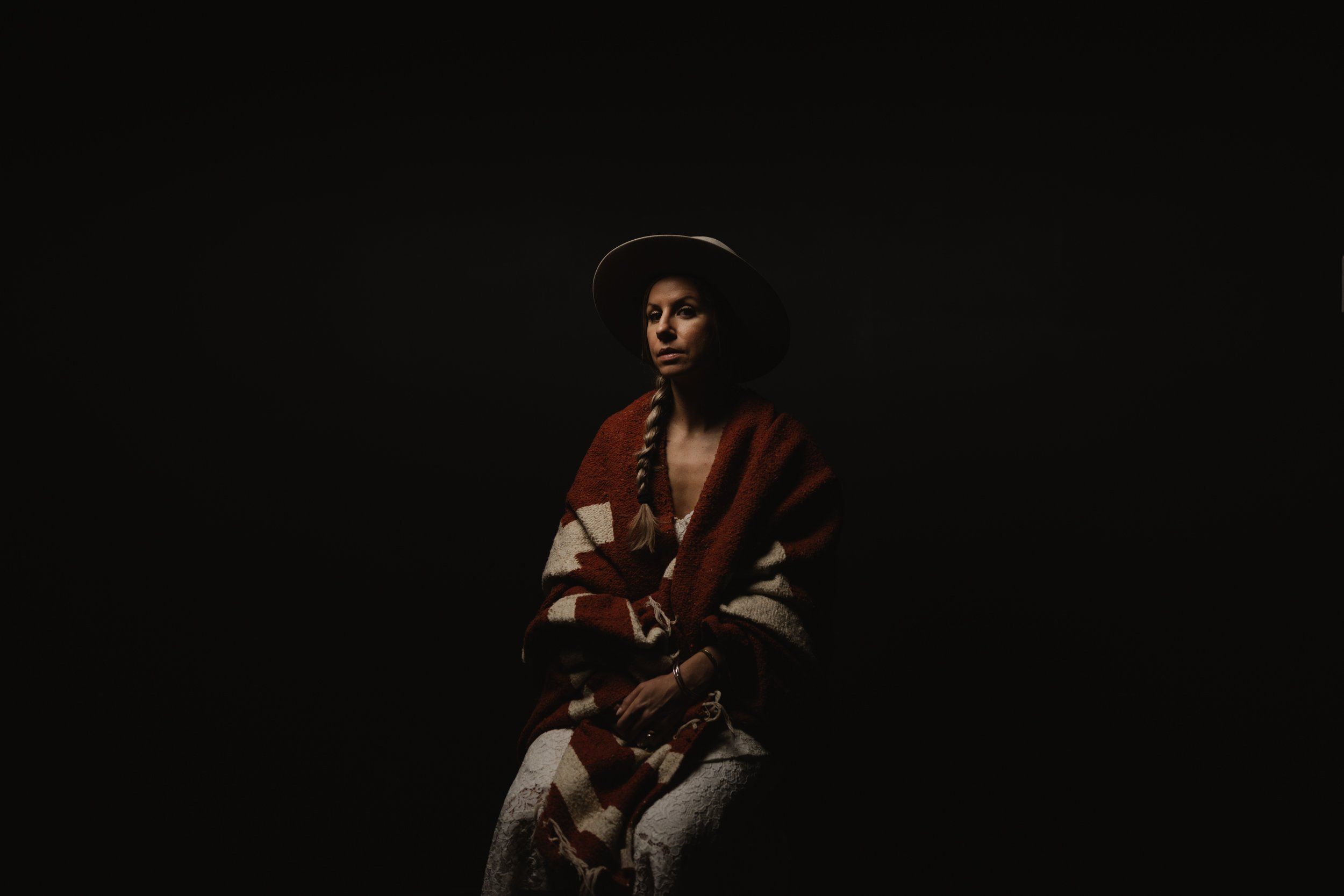Woman in burgundy and cream cardigan gazes downward against dark background, warm spotlight creating intimate portrait mood