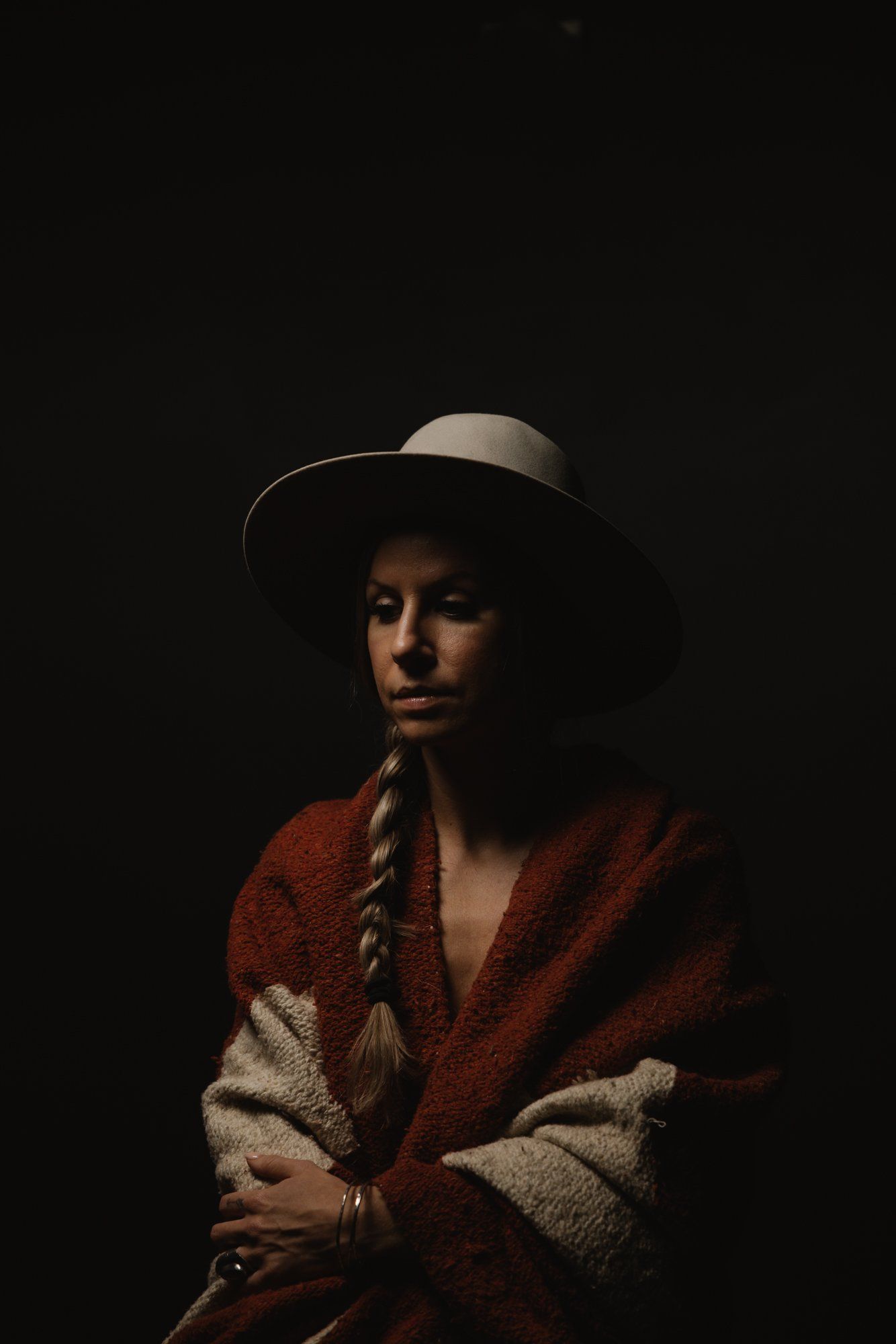 Woman in braided hair and rust-colored shawl gazes thoughtfully against dark background, wearing a wide-brimmed hat
