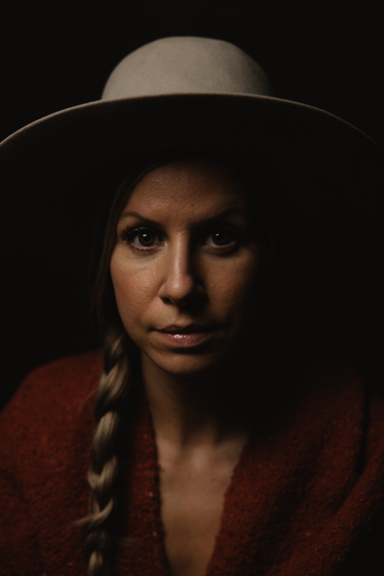Woman in red shirt and cream cowboy hat gazes directly at camera against dark background, braided hair visible at shoulder