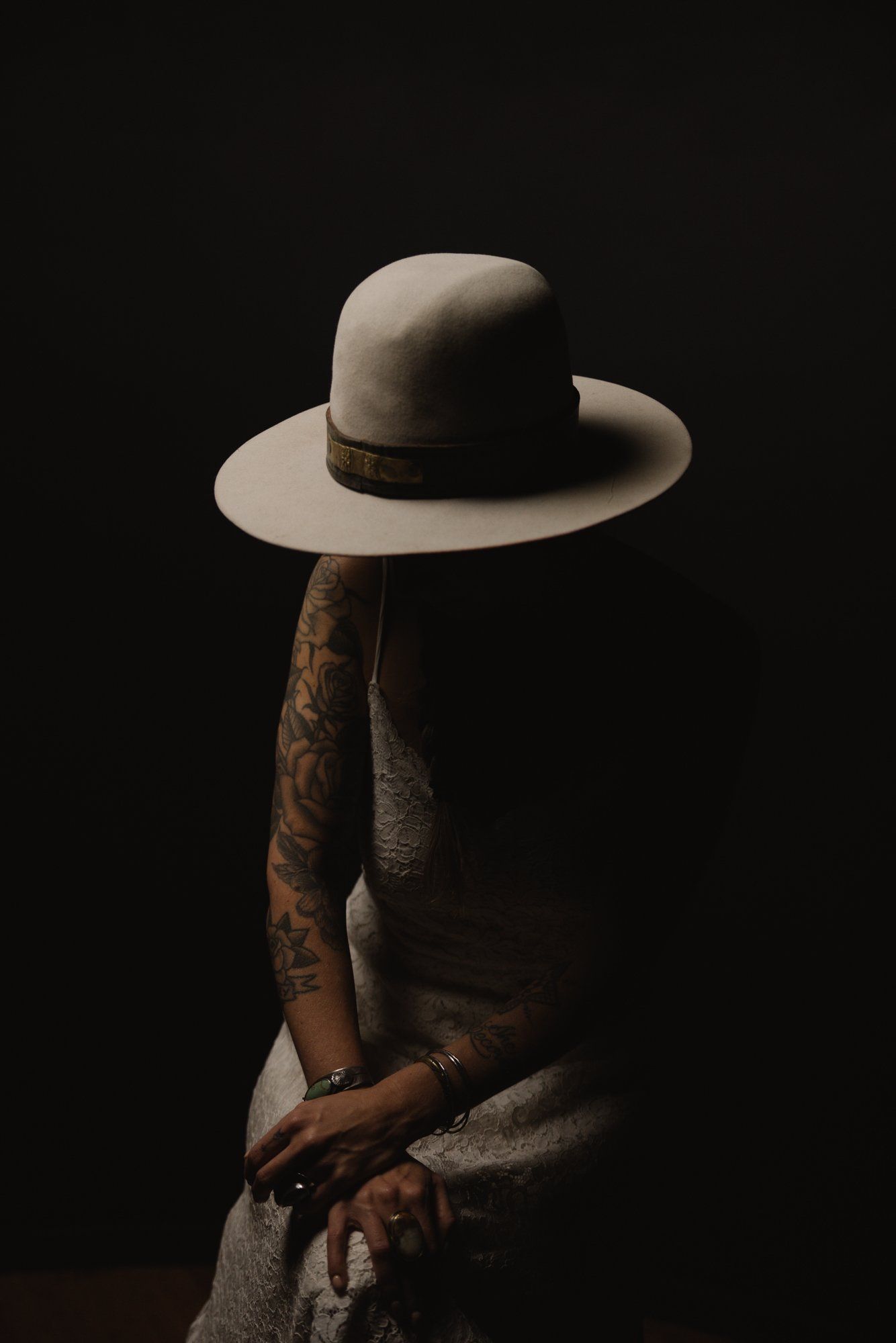 Tattooed man in light linen shirt holds cream-colored hat against dark background, dramatic side lighting casting deep shadows