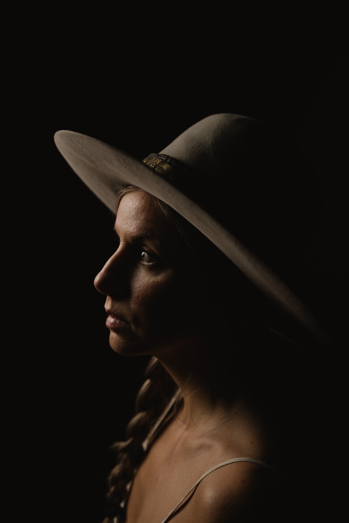 Profile portrait of woman wearing wide-brimmed hat against black background, dramatic side lighting illuminating her face and neck