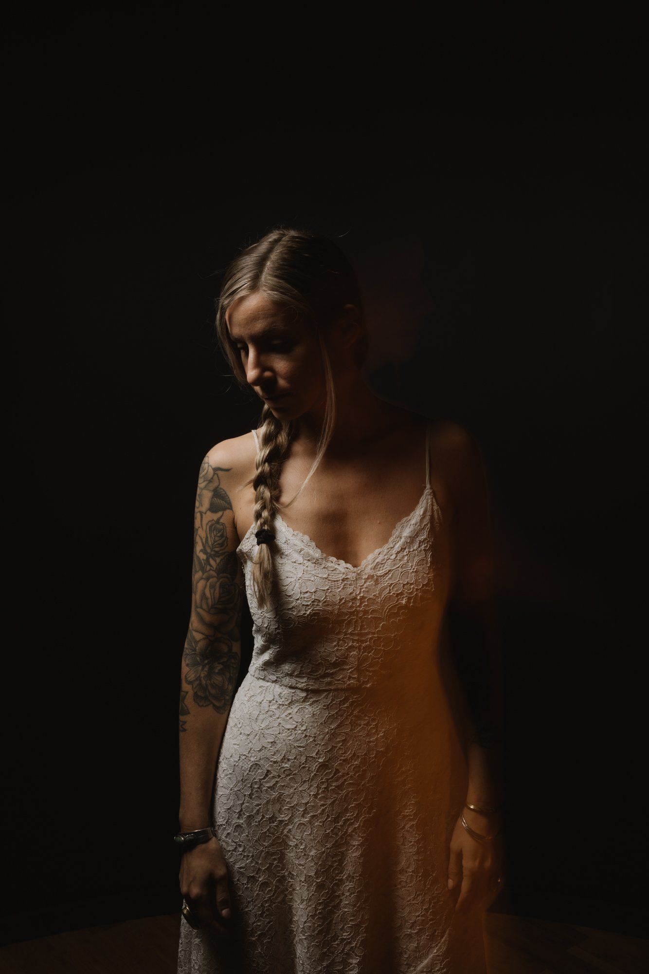 Woman in textured white dress with braided hair gazes downward against dramatic dark background, side-lit by warm golden light
