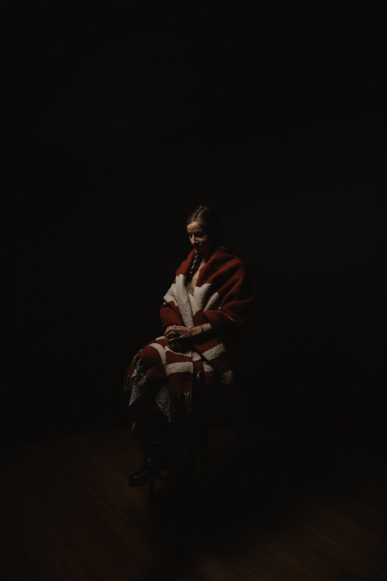 Woman in rust-colored jacket seated against deep black background, warm spotlight illuminating her contemplative expression and clasped hands