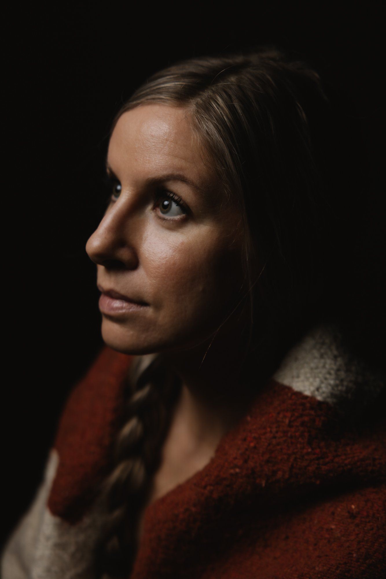 Woman with braided hair wearing a deep red cardigan, photographed in profile against a dark background with warm, directional lighting