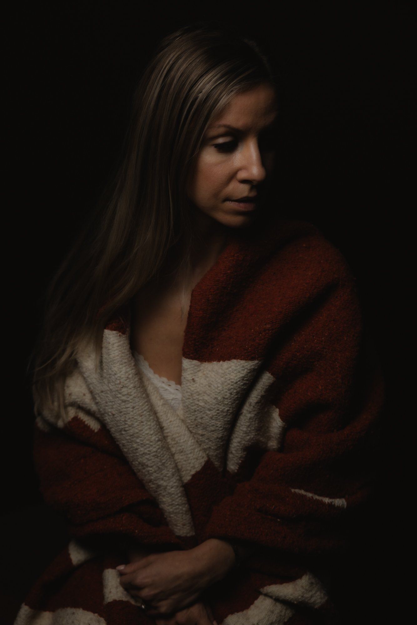 Woman with long dark hair wearing a rust and cream cardigan gazes downward against a dark background in soft, dramatic studio lighting