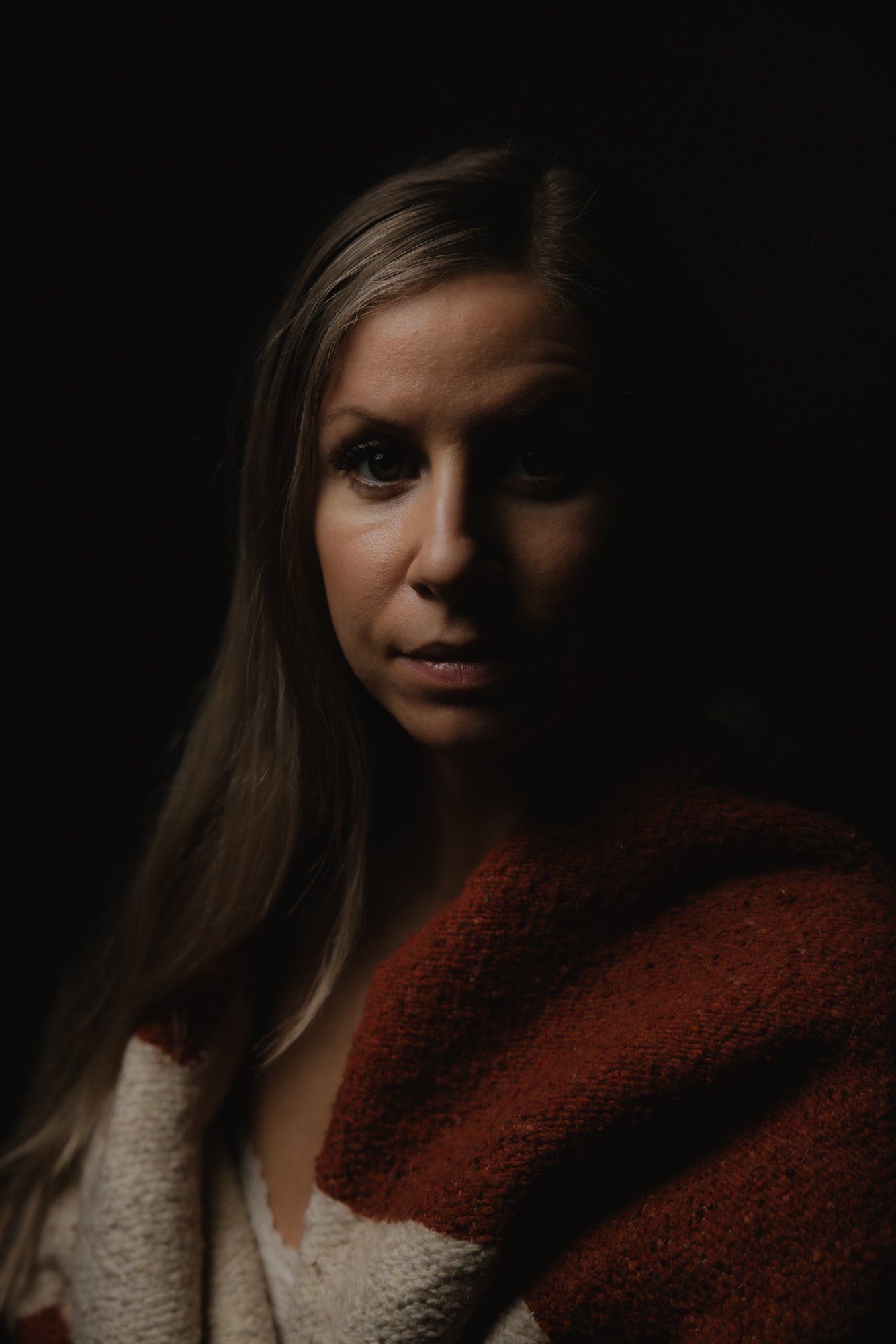 Woman with long brown hair wearing a rust and cream layered garment, photographed against black background with dramatic side lighting