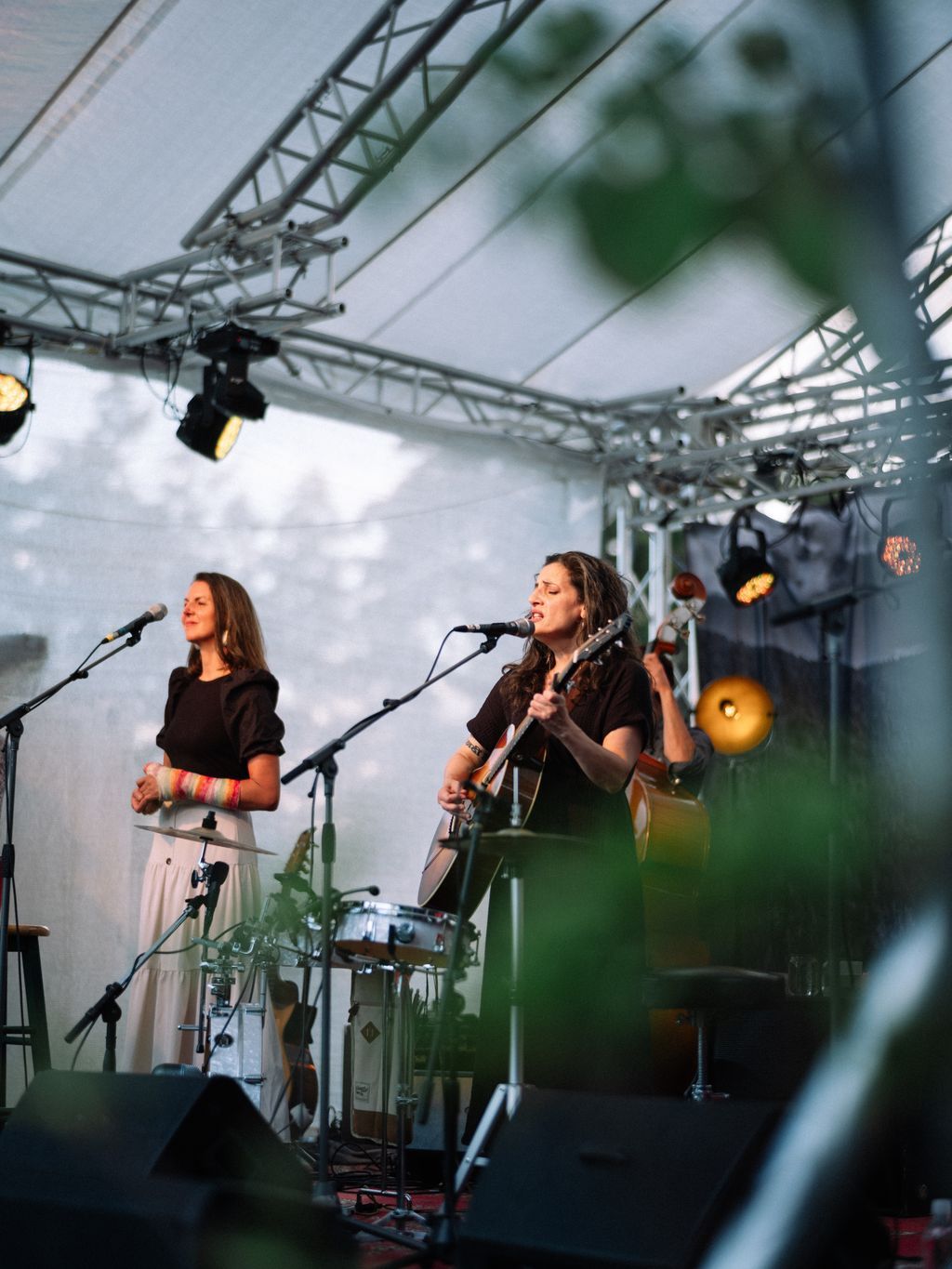Musician performs with acoustic guitar at outdoor stage under metal truss framework, cloudy sky behind