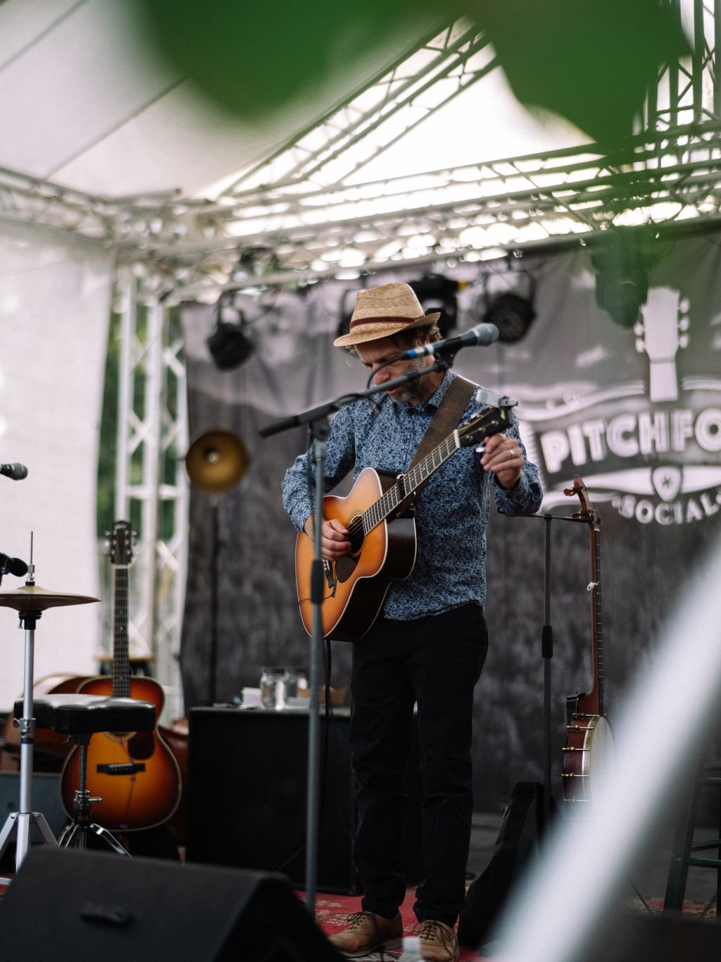 Musician in straw hat and blue shirt plays acoustic guitar at microphone under greenhouse roof panels on Salt Spring Island