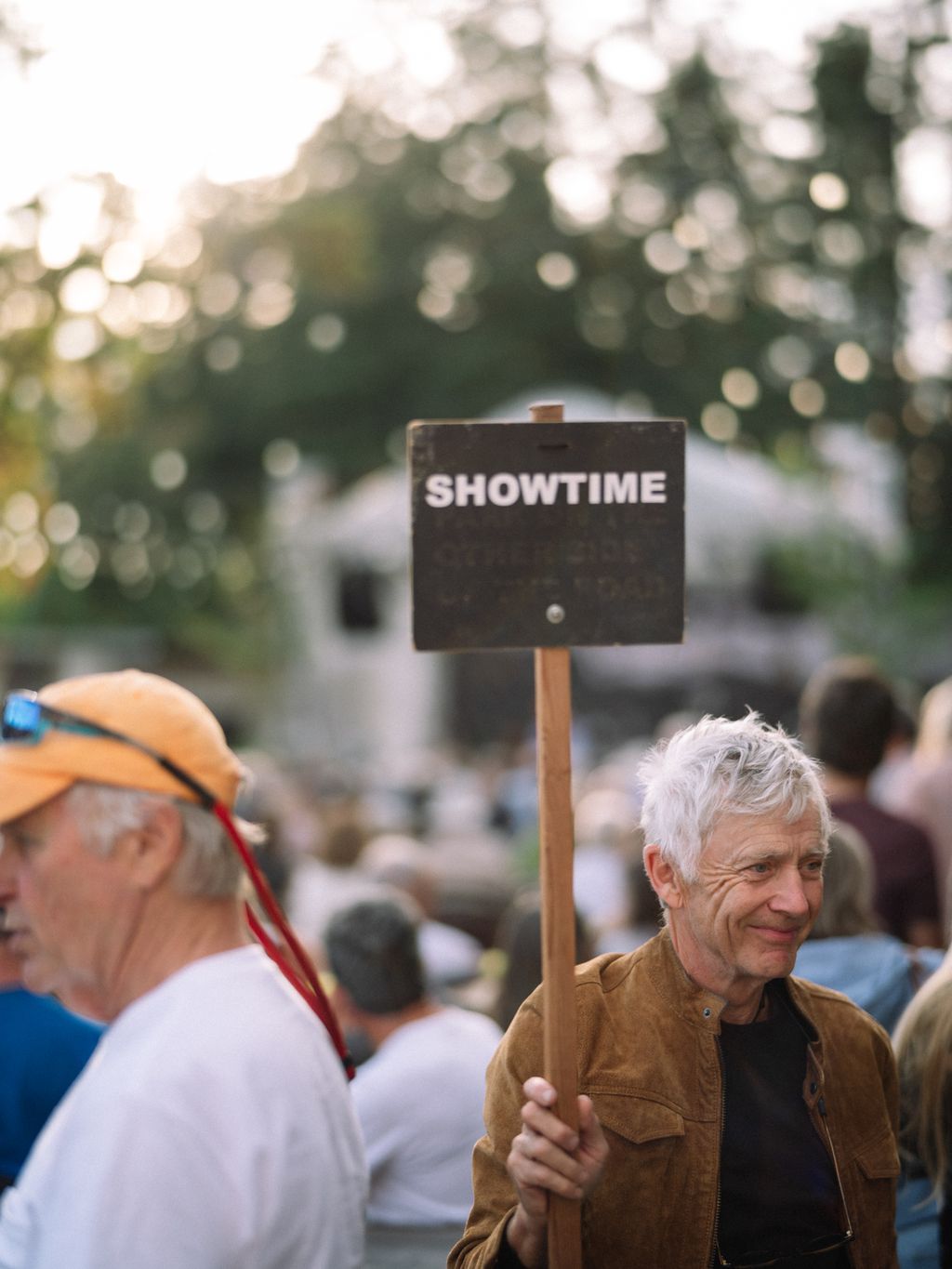 Man holding "Showtime" sign at outdoor event with blurred crowd and flowering branches overhead on Salt Spring Island