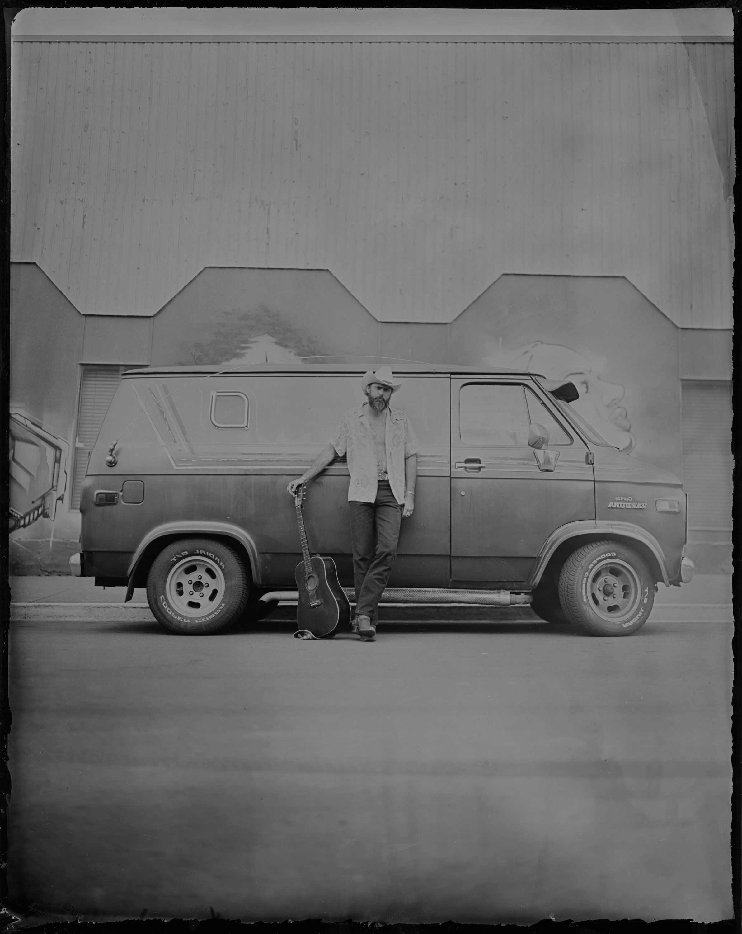 Woman in white shirt leaning against vintage van under geometric concrete overhang, black and white tintype style photograph