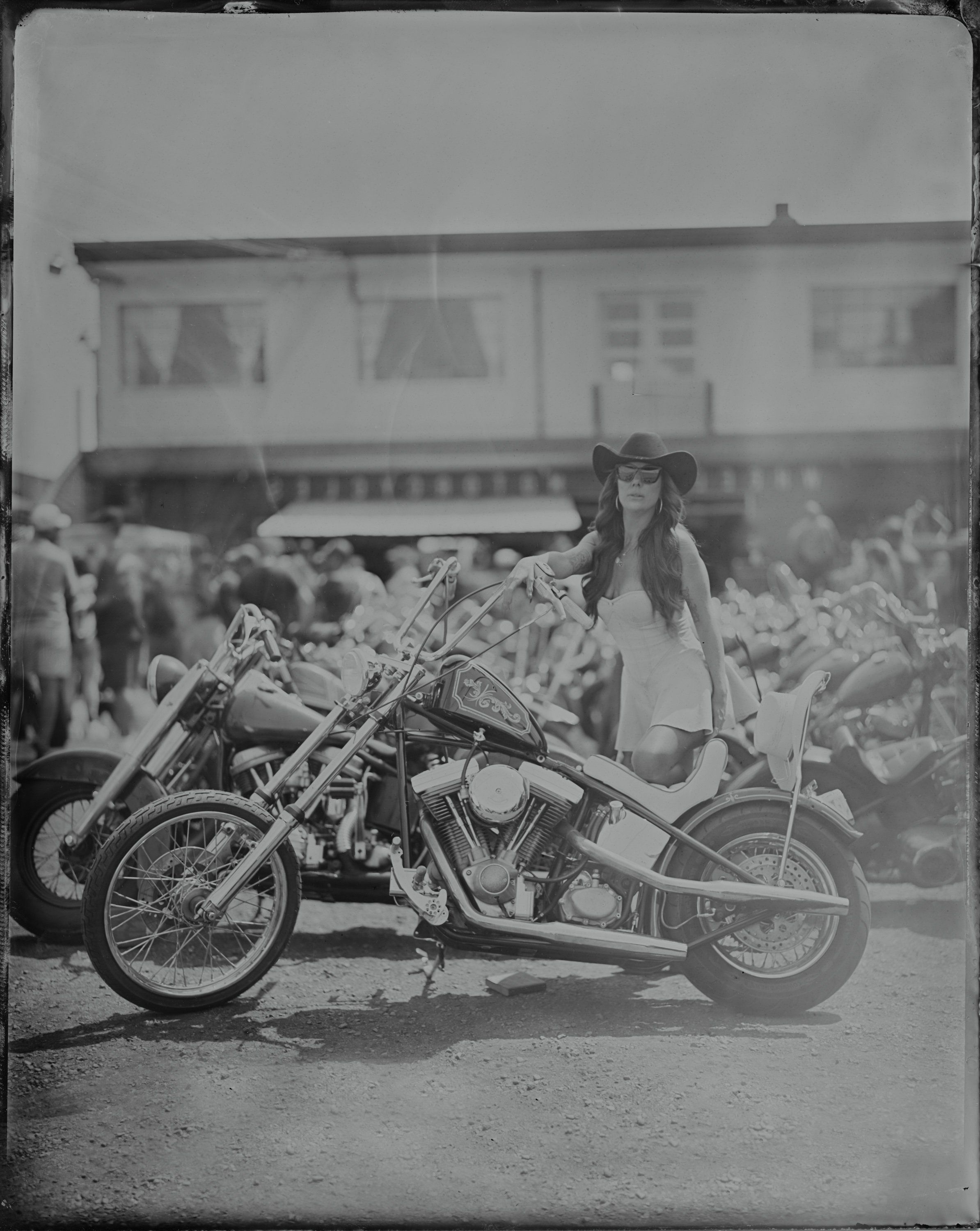 Woman in vintage dress seated on ornate chopper motorcycle, crowd and trailer visible behind in parking lot celebration