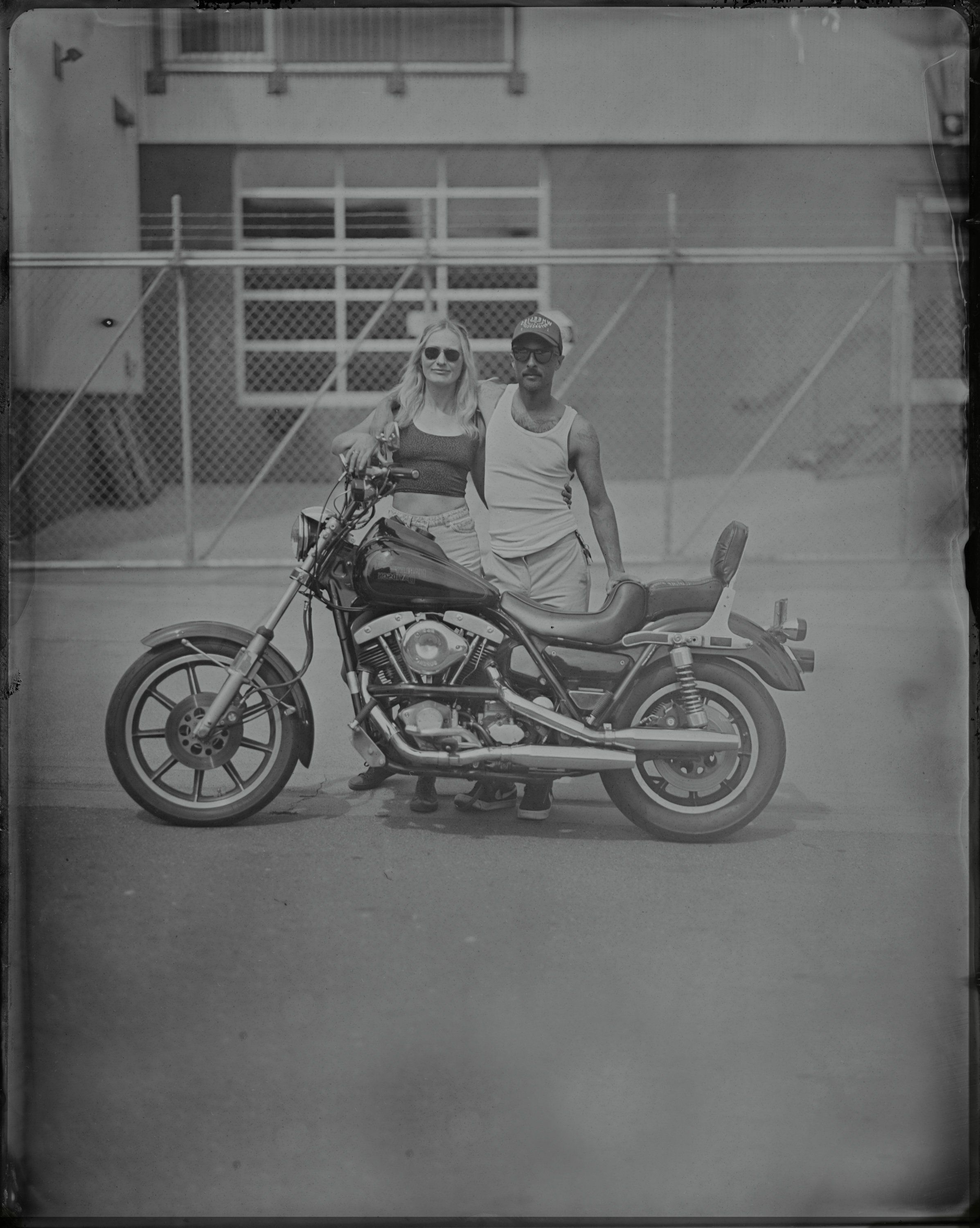 Two people sit on a vintage motorcycle in a parking lot, posed against a chain-link fence under bright sunlight, captured in black and white